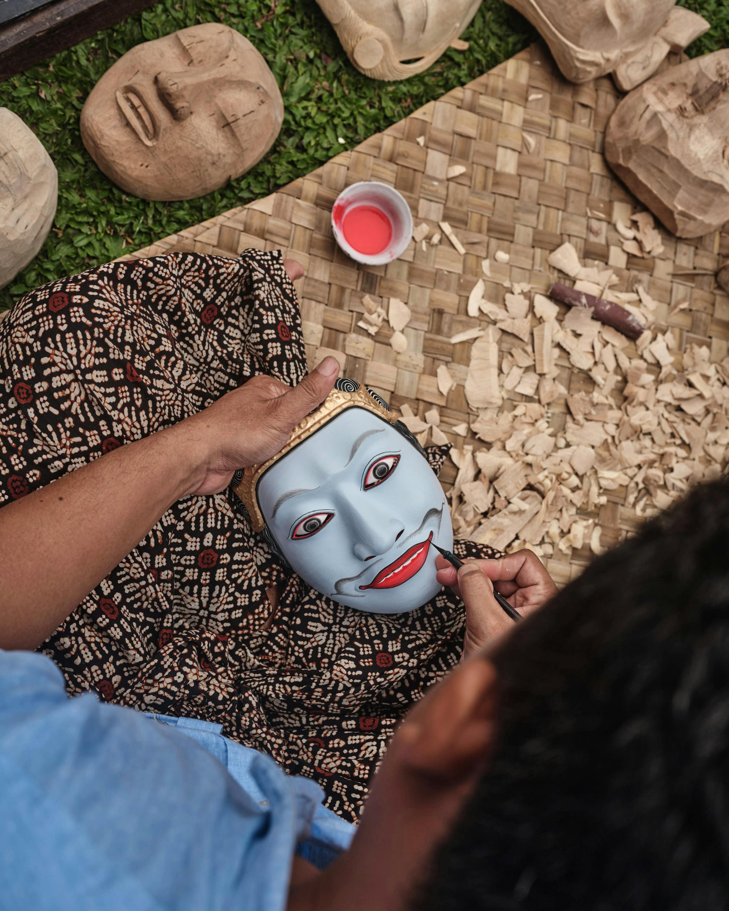 Hands paint a blue mask on a woven mat with art supplies nearby.