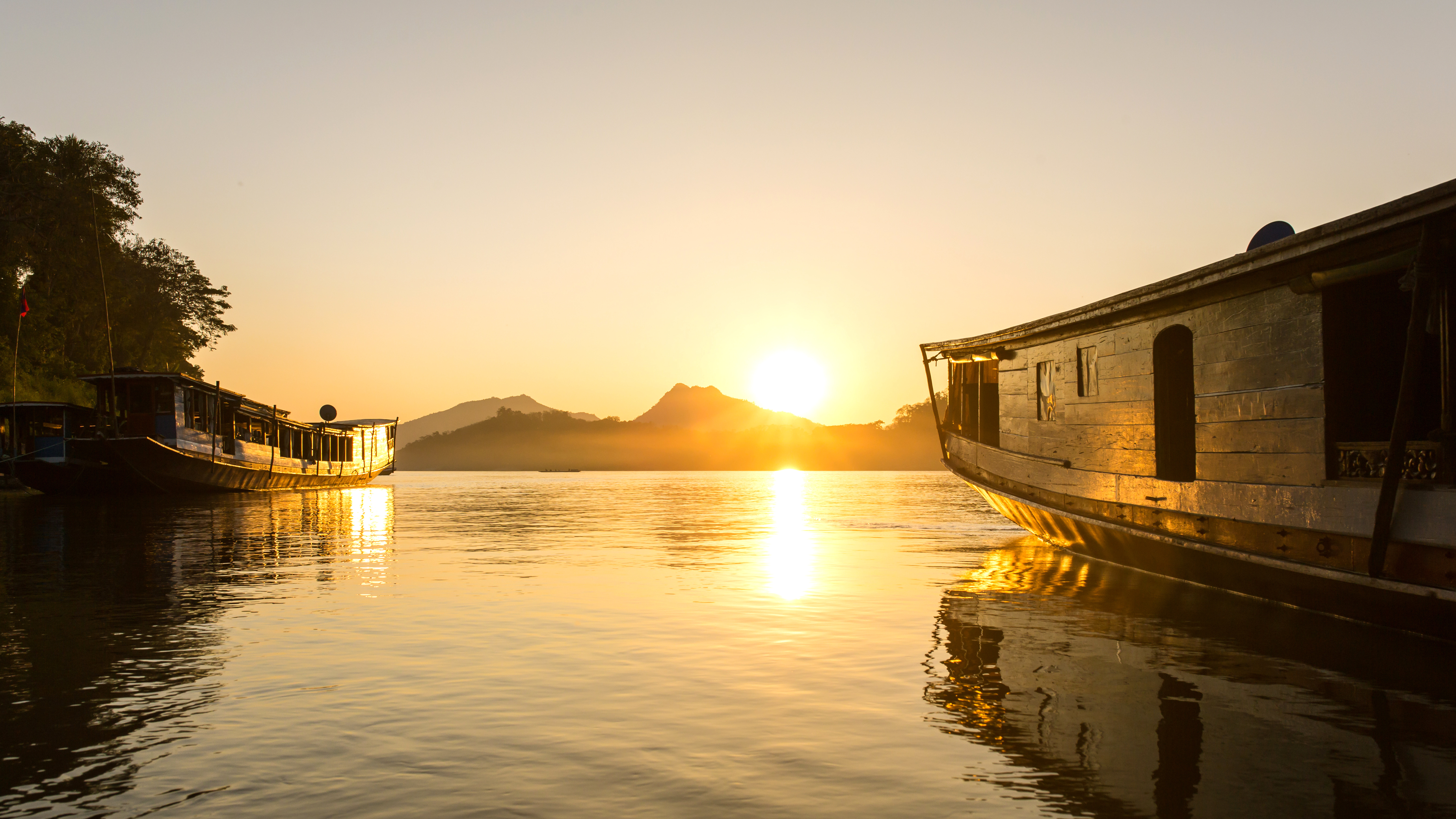 Sunset over a calm river with long boats and distant hills.