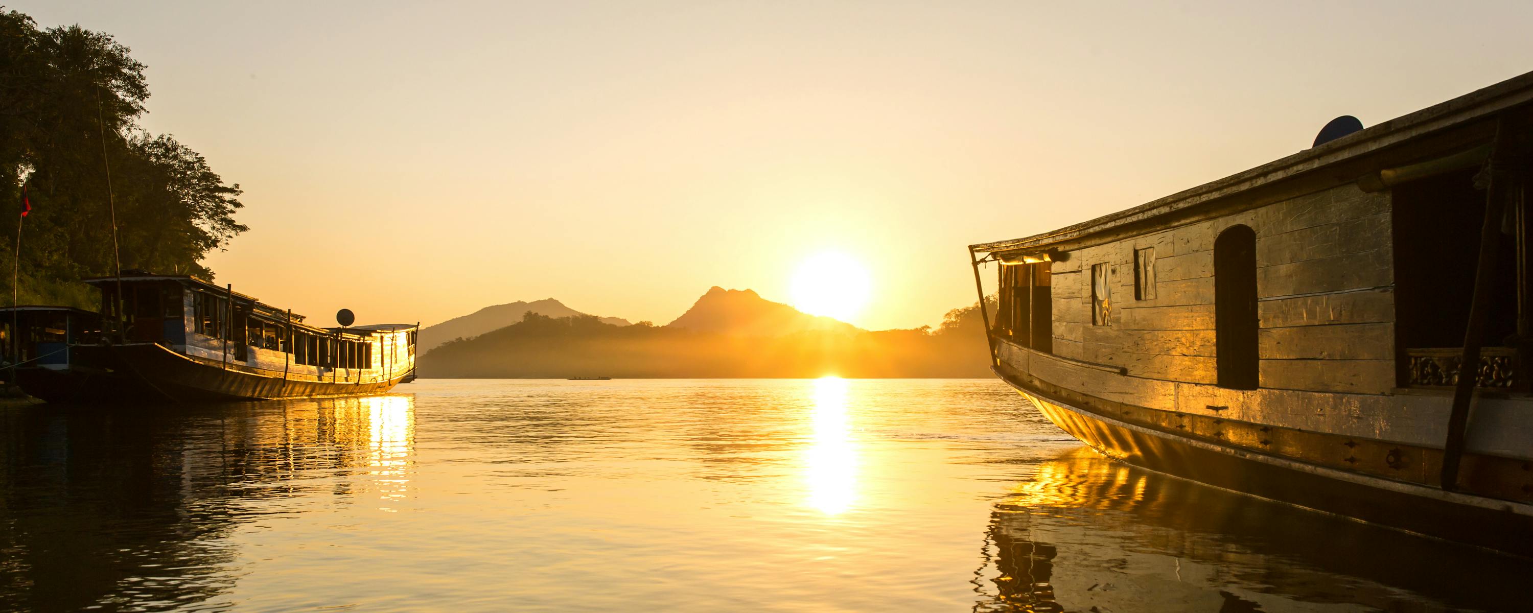 Sunset over a calm river with long boats and distant hills.