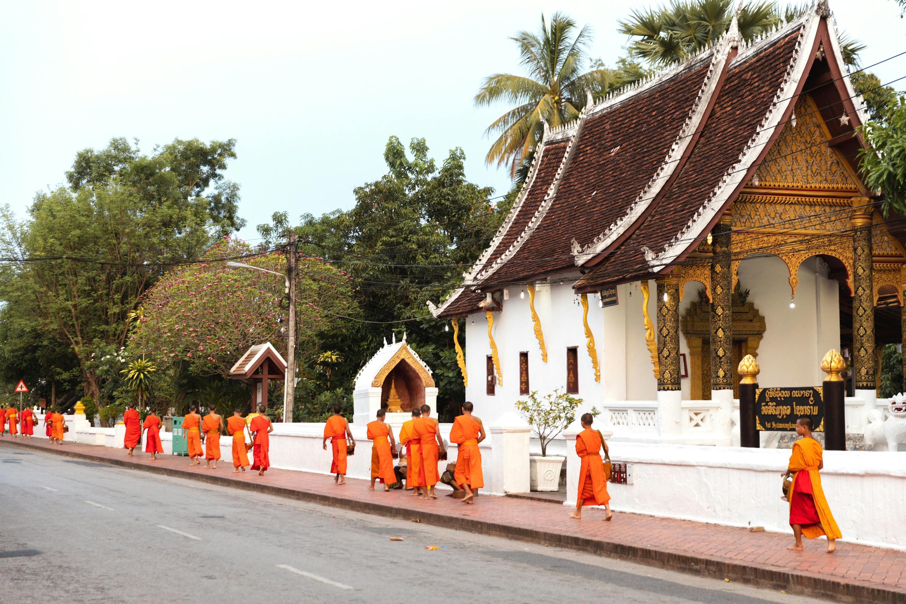 People in orange robes walk along a street beside a white temple wall.