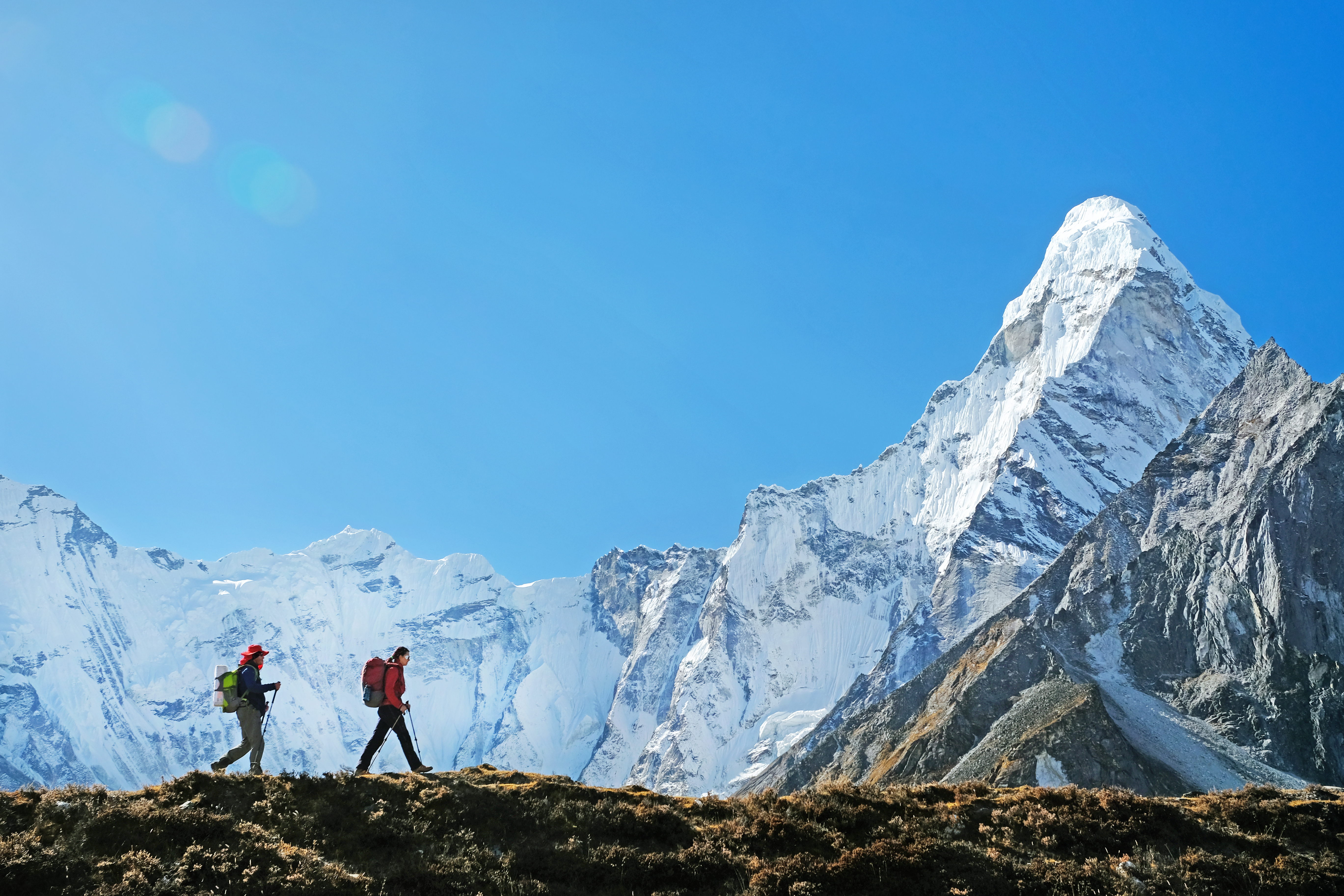 Two hikers walk along a grassy ridge with towering snowy peaks behind them.