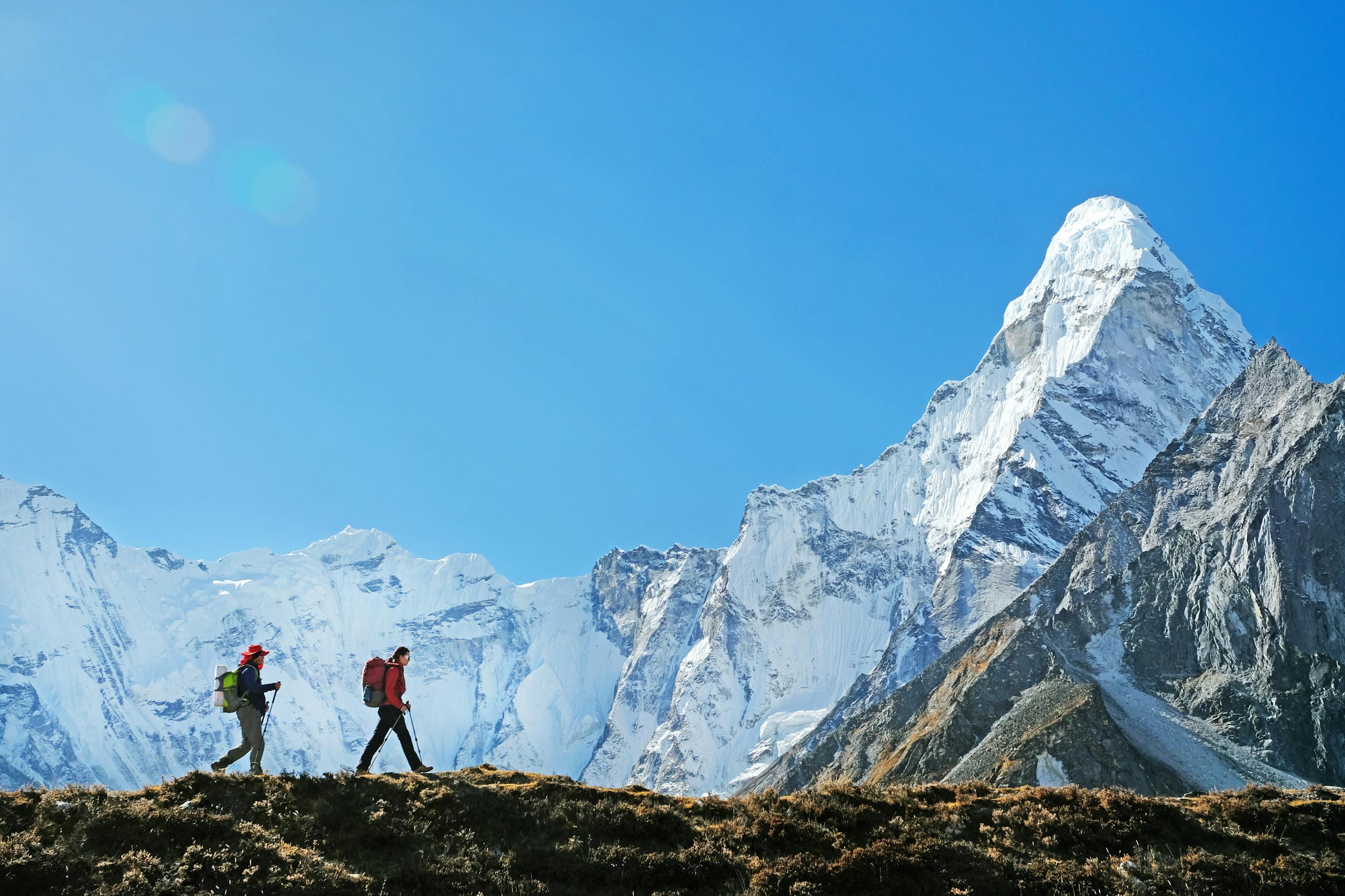 Two hikers walk along a grassy ridge with towering snowy peaks behind them.