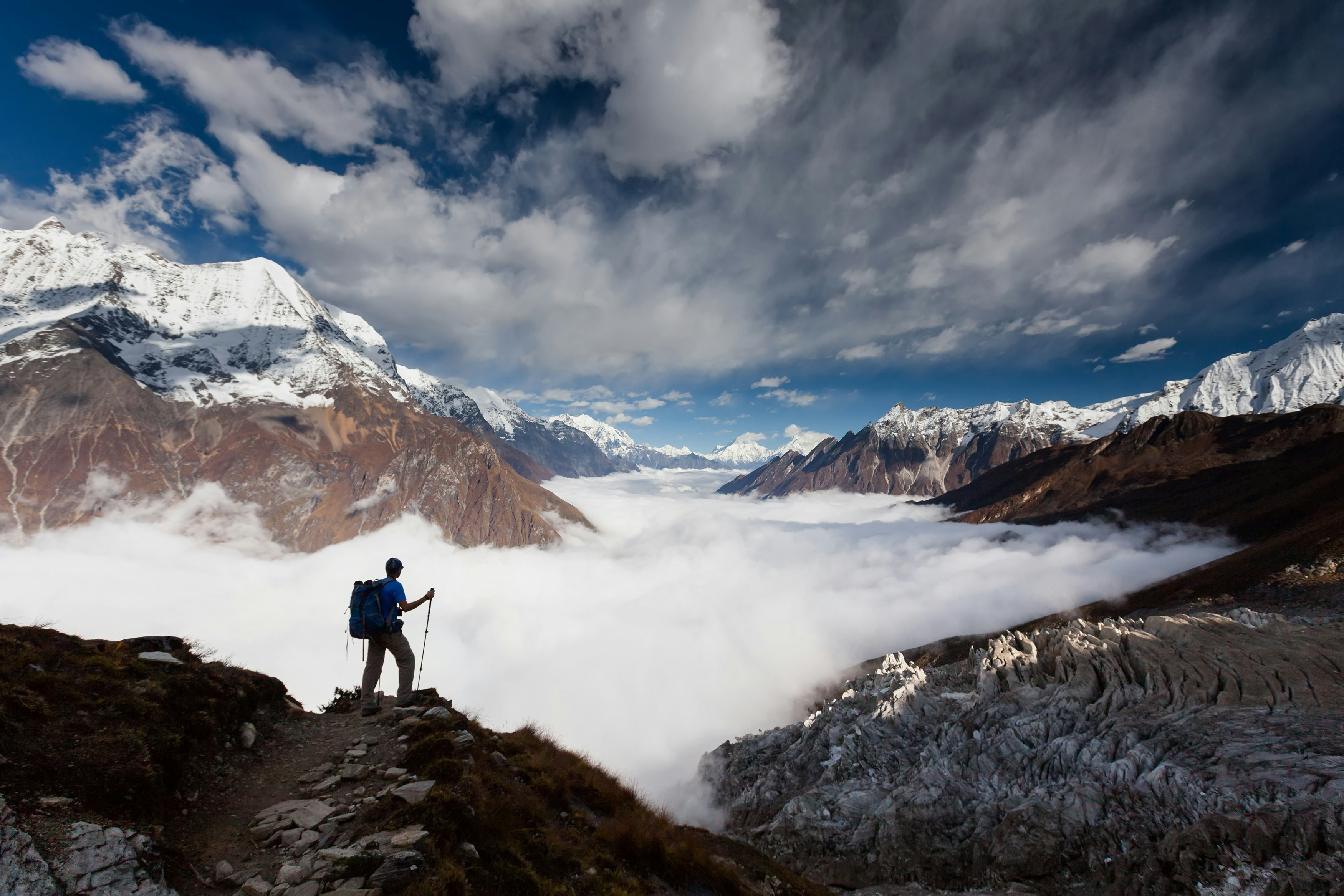 Person stands on a ridge above clouds with snowcapped mountains under a dramatic sky.