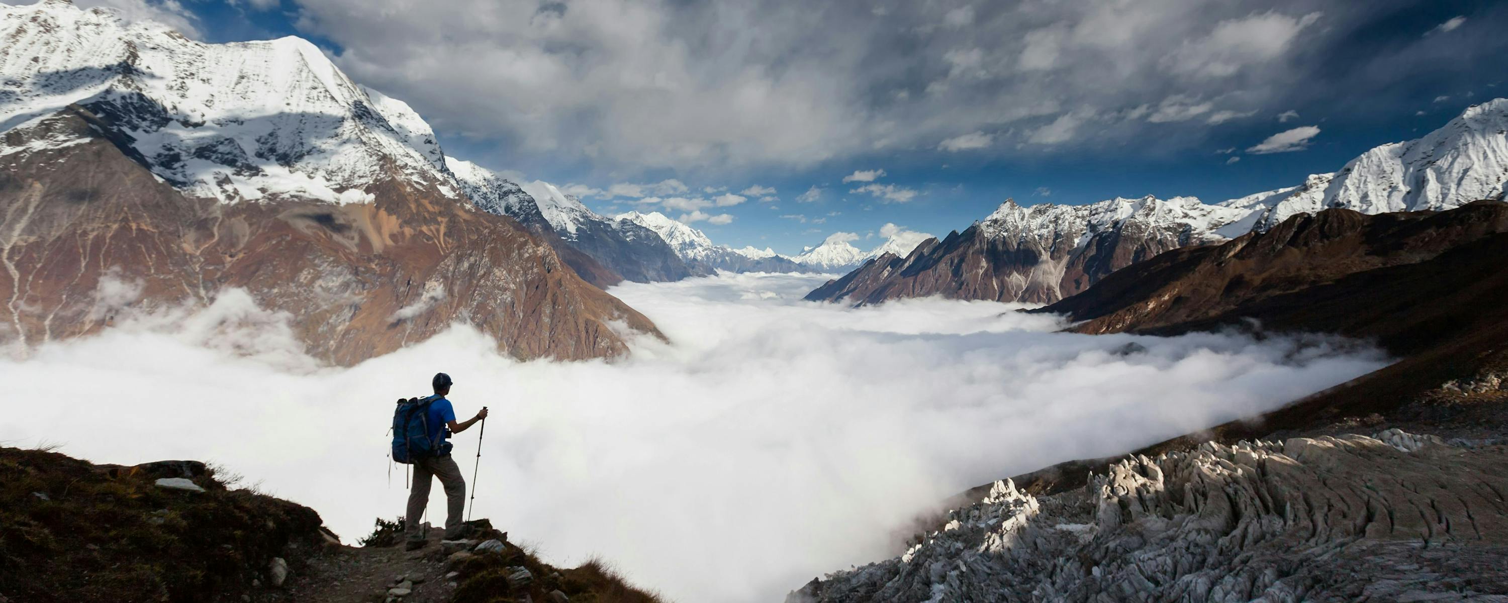 Person stands on a ridge above clouds with snowcapped mountains under a dramatic sky.