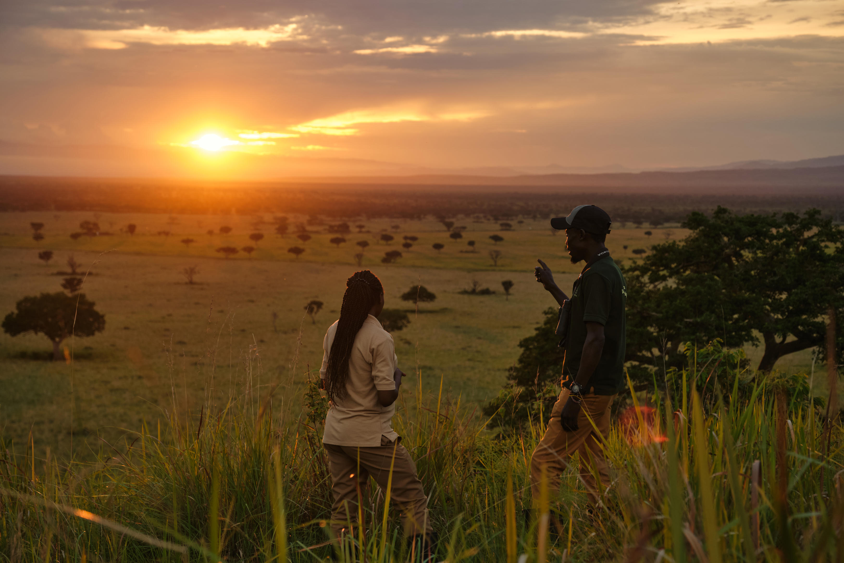 Two travelers stand in tall grass at sunset, overlooking a wide savanna with scattered trees and low haze.