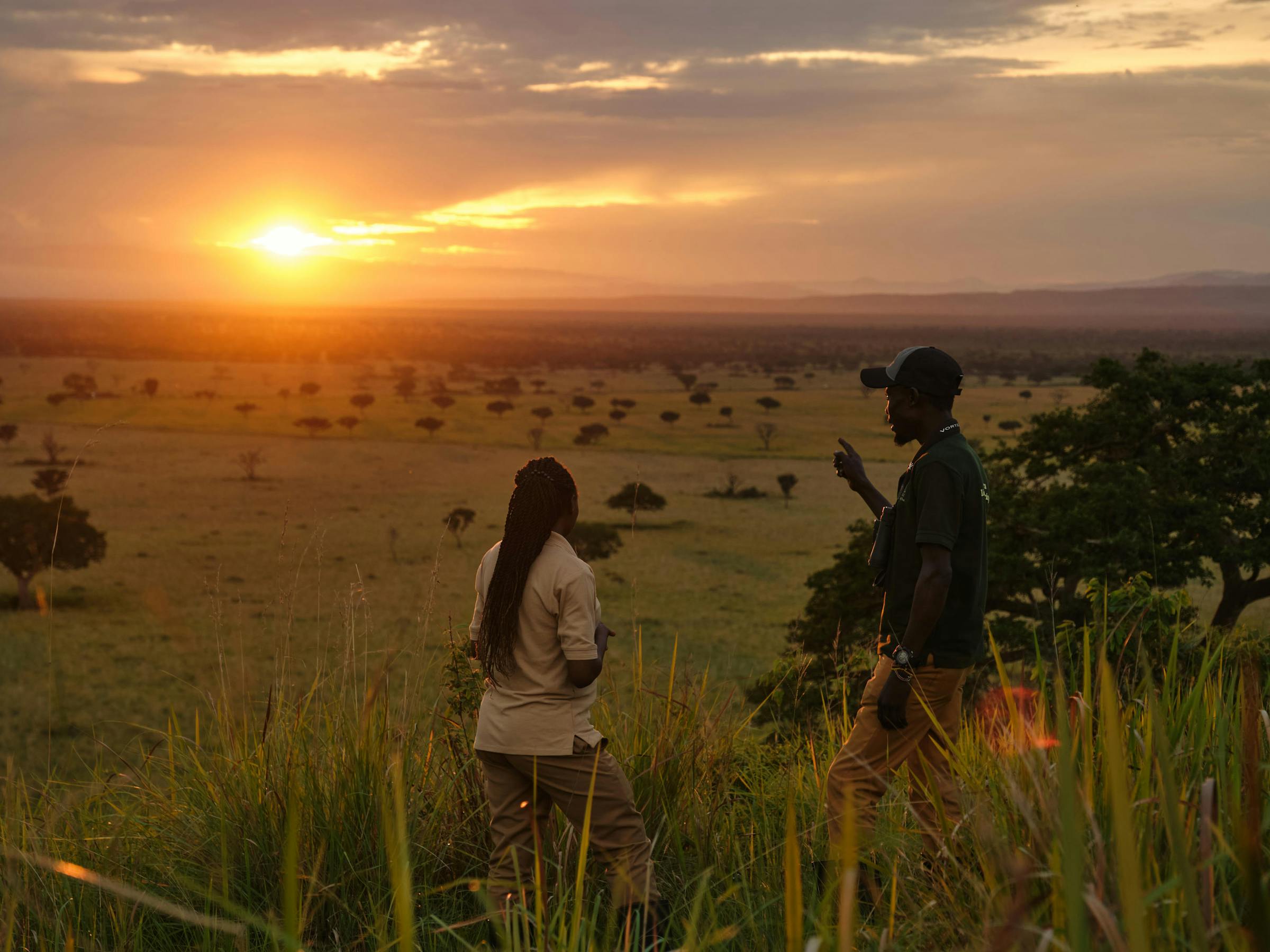 Two travelers stand in tall grass at sunset, overlooking a wide savanna with scattered trees and low haze.