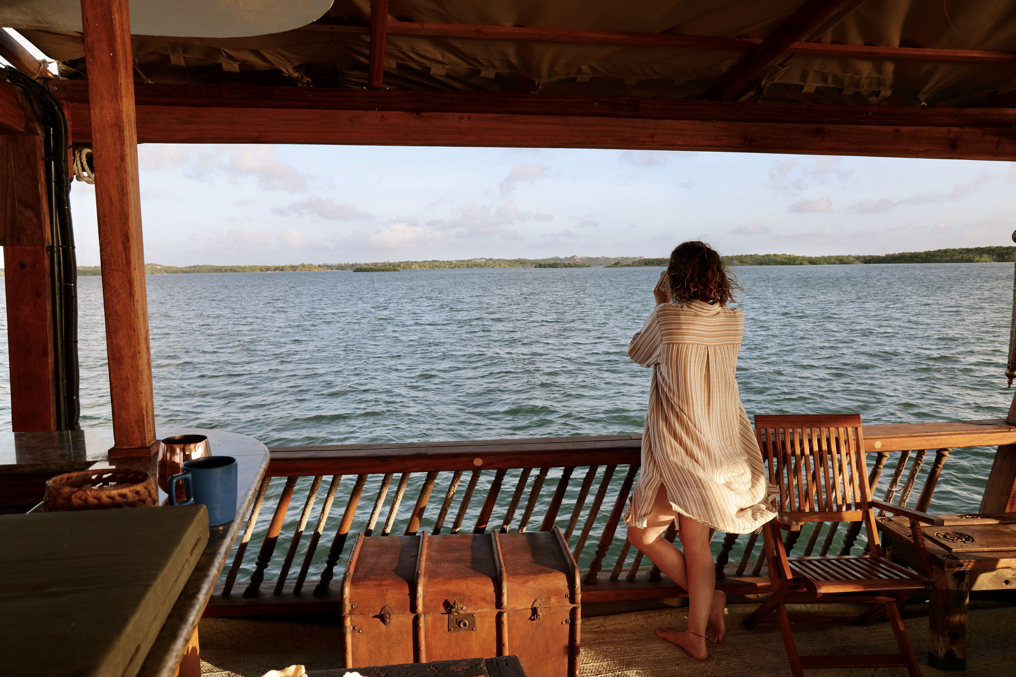 A person stands on a shaded wooden deck facing calm water, with railings and distant shoreline under a clear sky.