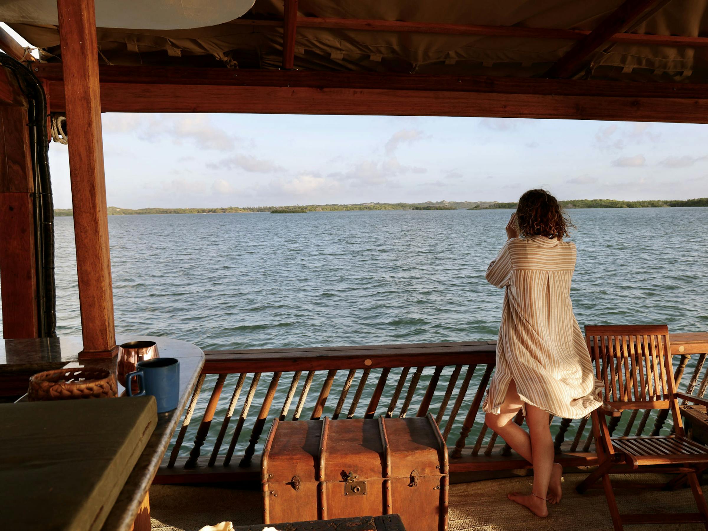 A person stands on a shaded wooden deck facing calm water, with railings and distant shoreline under a clear sky.