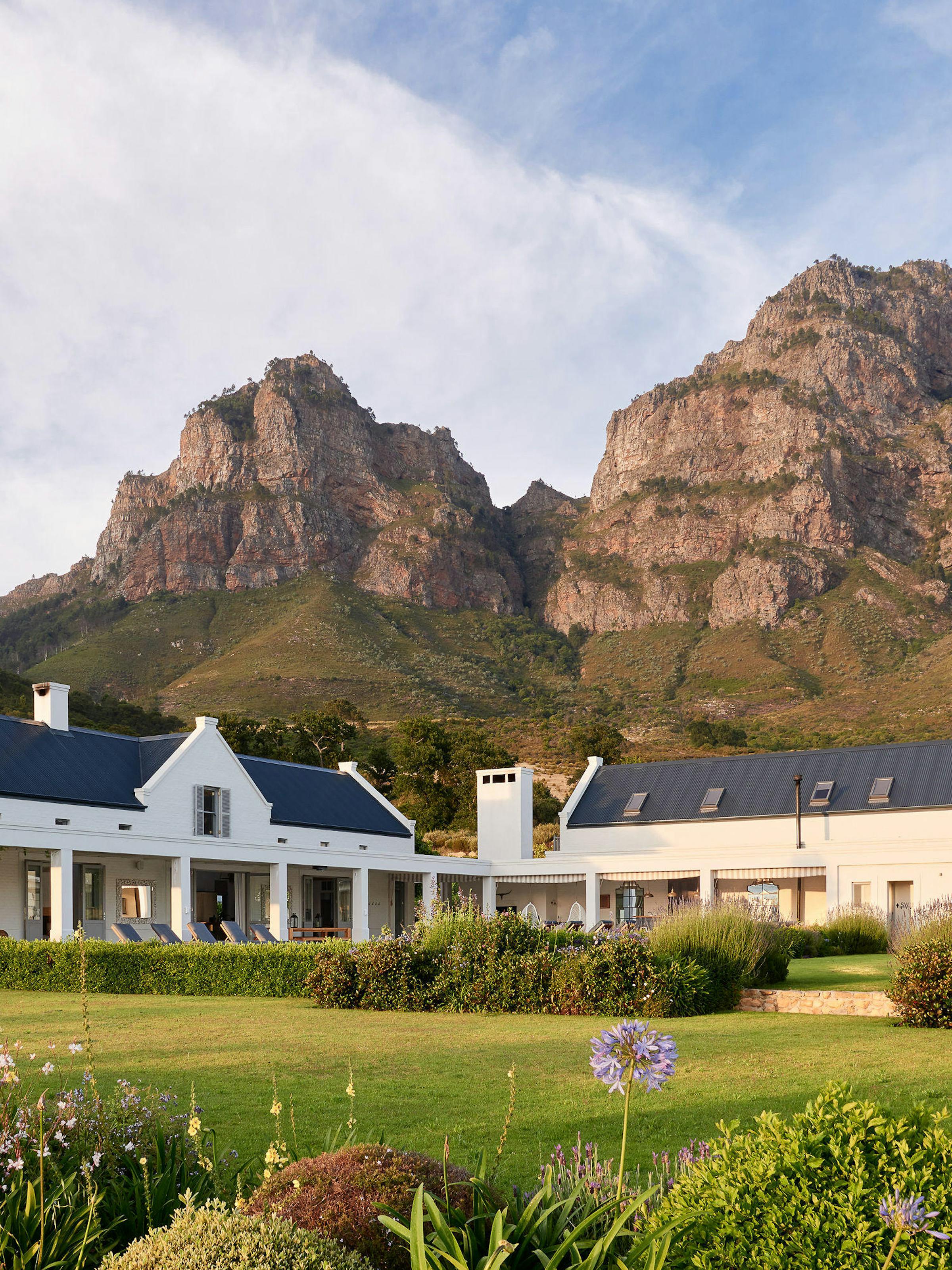 A white country villa sits beneath rugged mountains, surrounded by lawns, flowers, and tall trees in warm light.