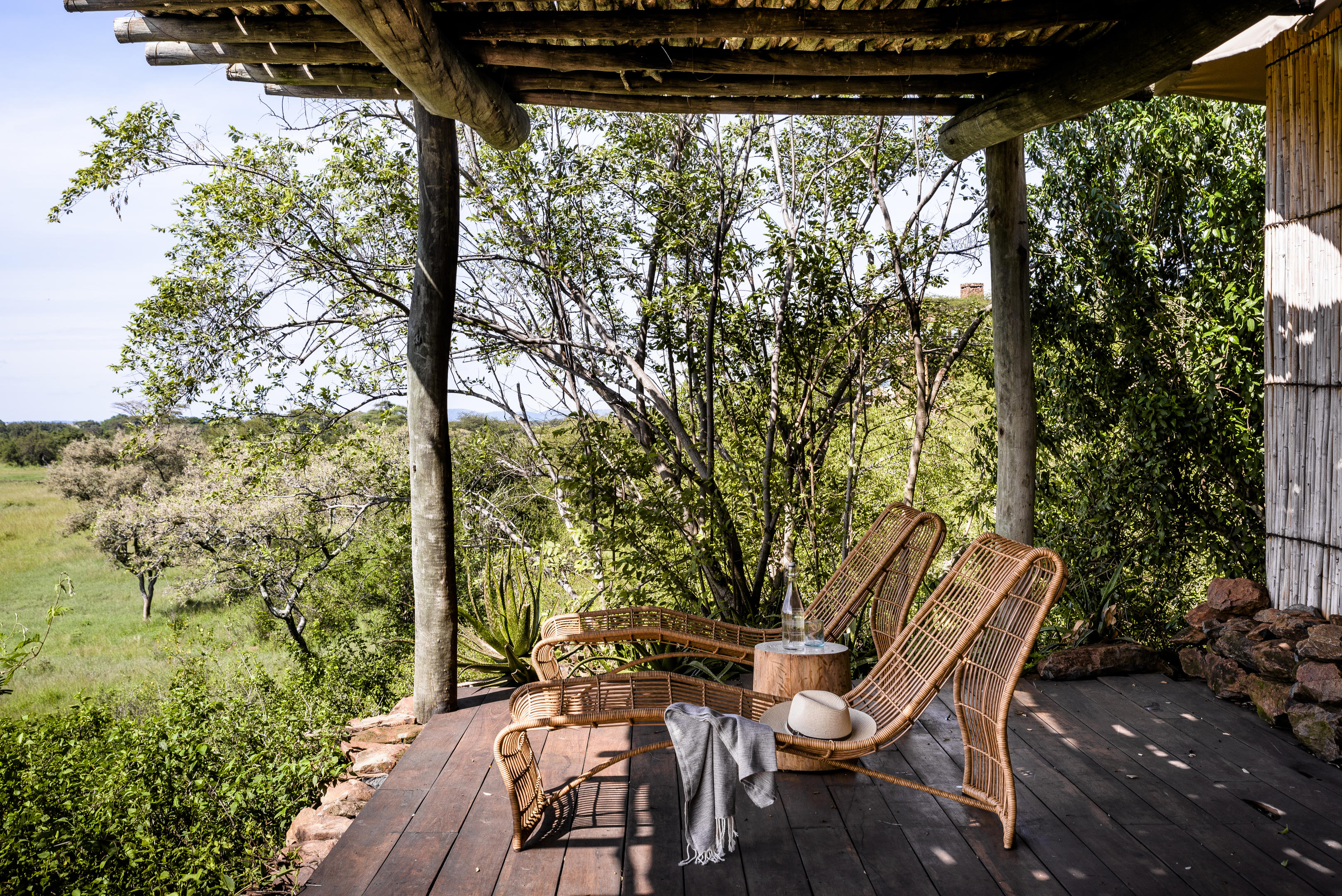 Wooden deck with chairs and a canvas canopy overlooks grassy plains at sunrise, with a lone acacia tree beyond.