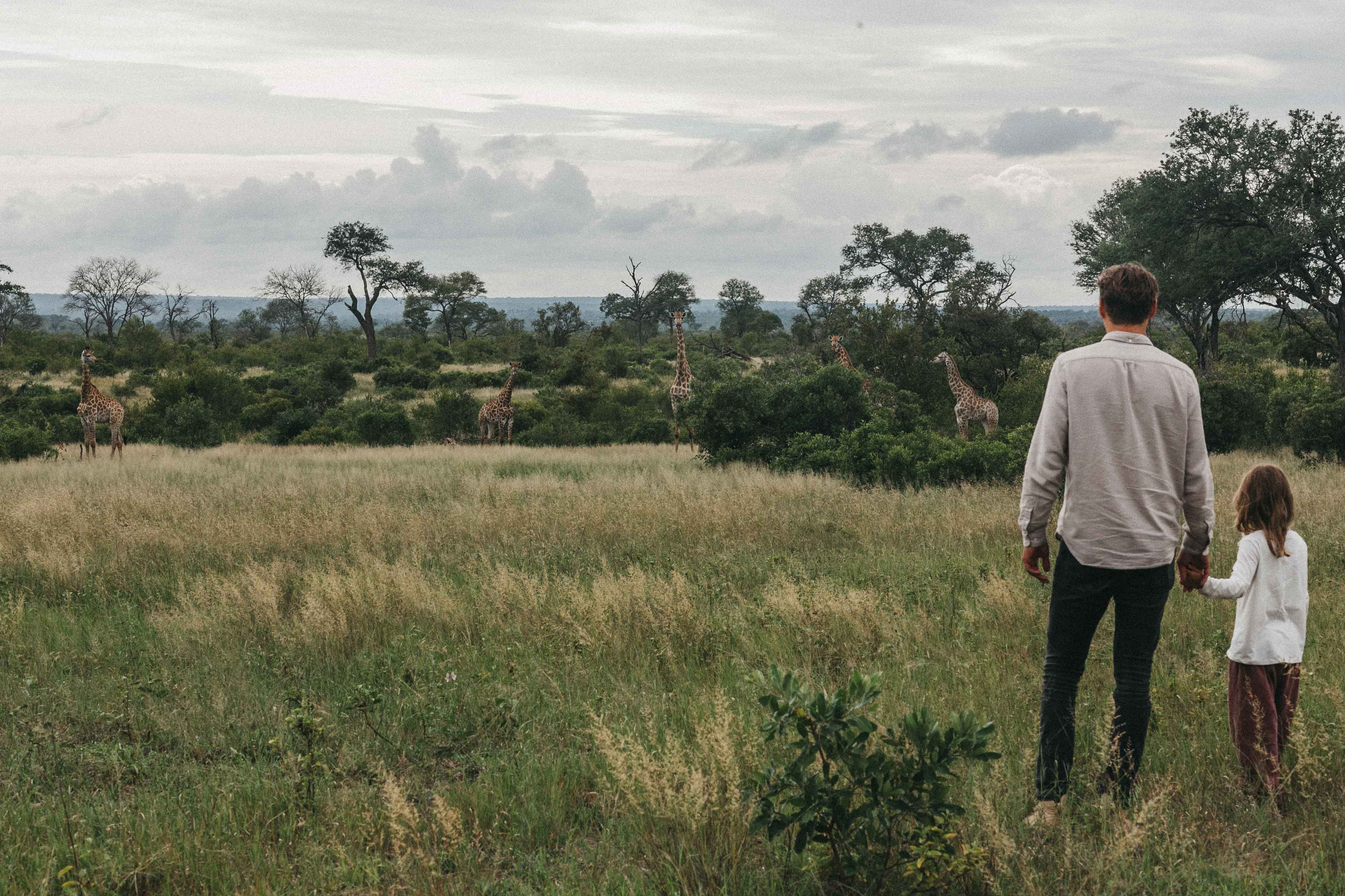 An adult and a child walk through tall grass toward distant trees under a cloudy sky across a wide savanna.