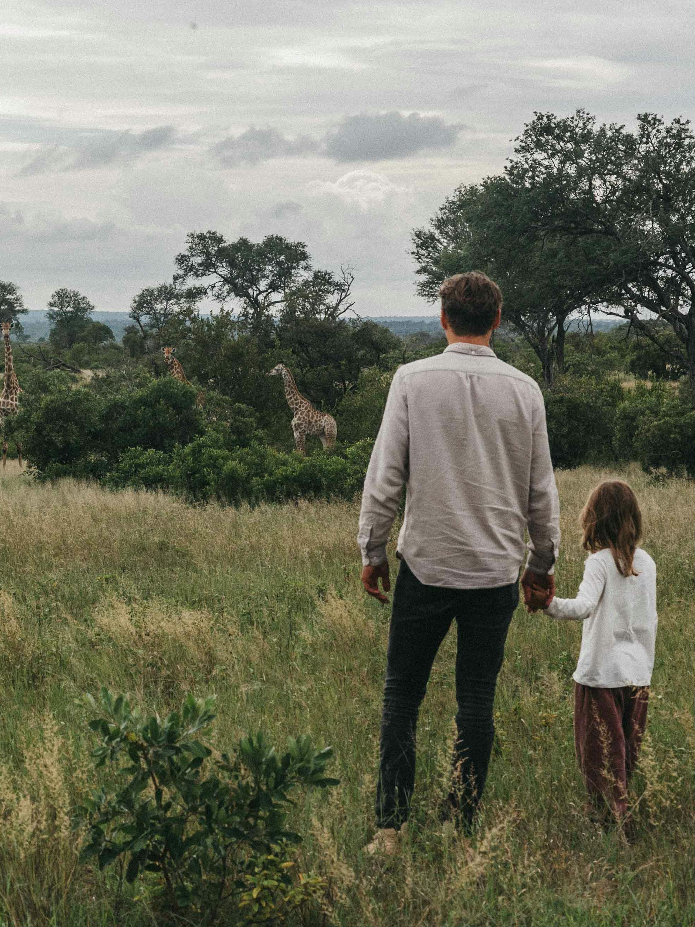 An adult and a child walk through tall grass toward distant trees under a cloudy sky across a wide savanna.