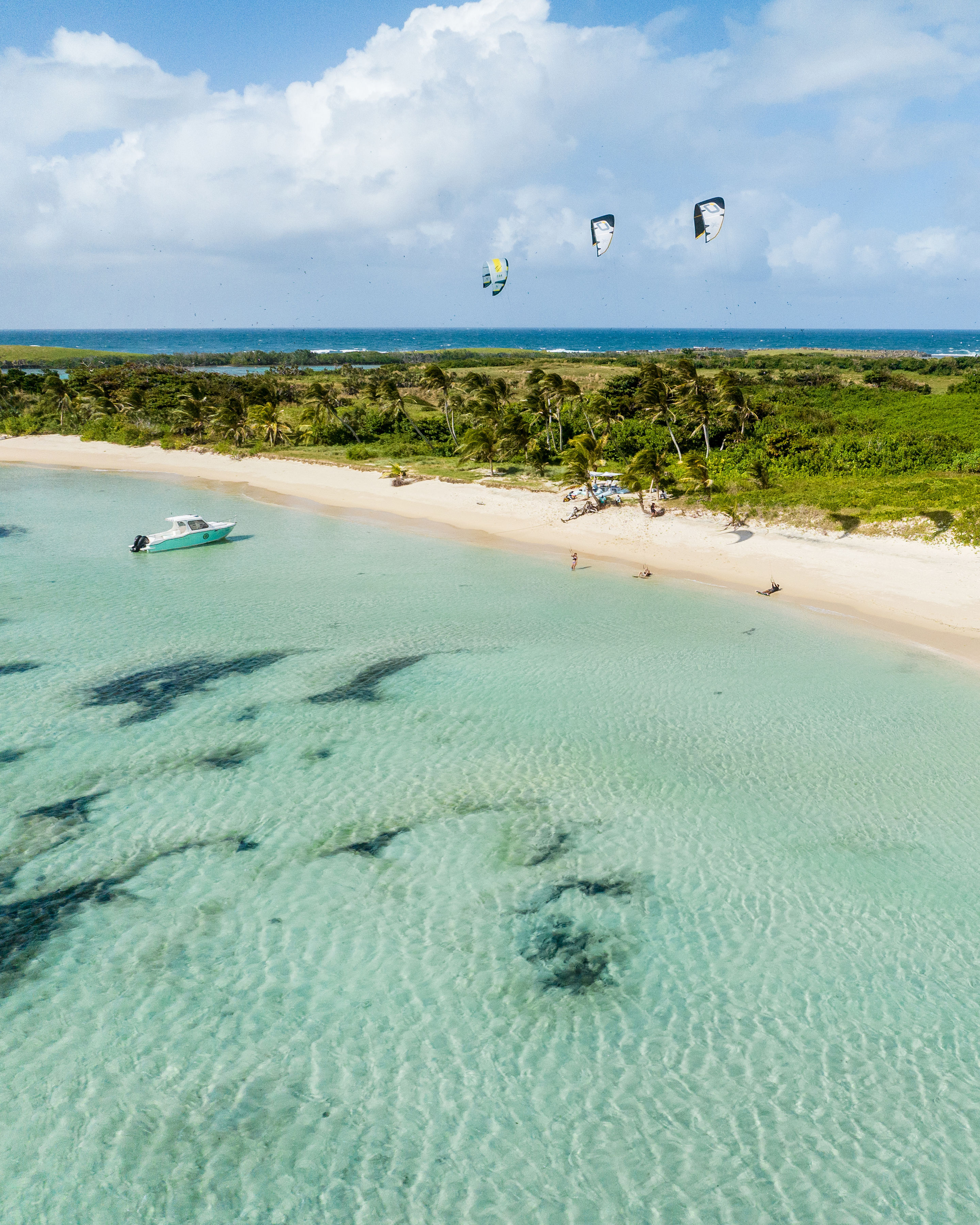 Aerial view of a white-sand beach and turquoise lagoon beside a lush island, with kitesurfers in the shallows.