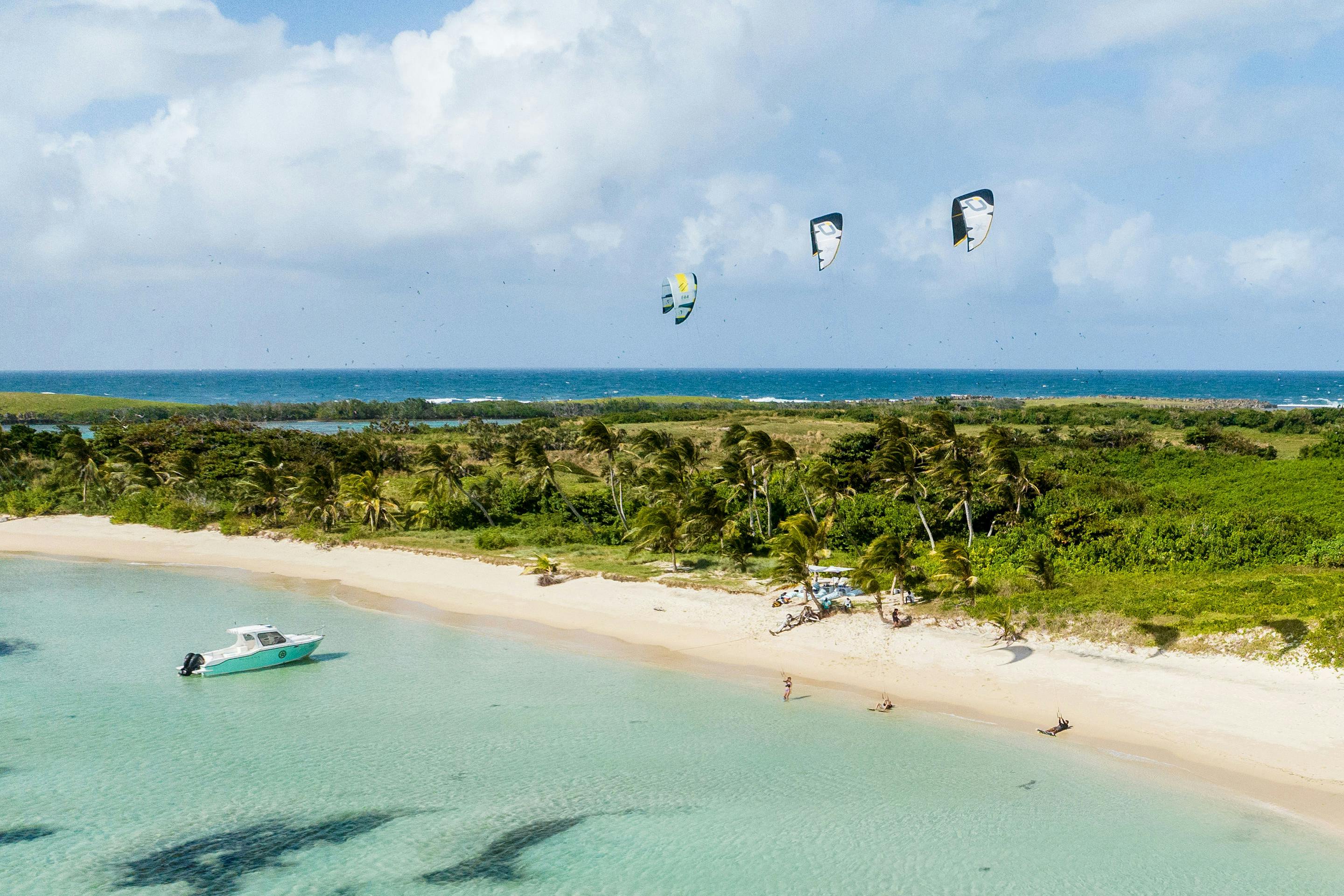 Aerial view of a white-sand beach and turquoise lagoon beside a lush island, with kitesurfers in the shallows.