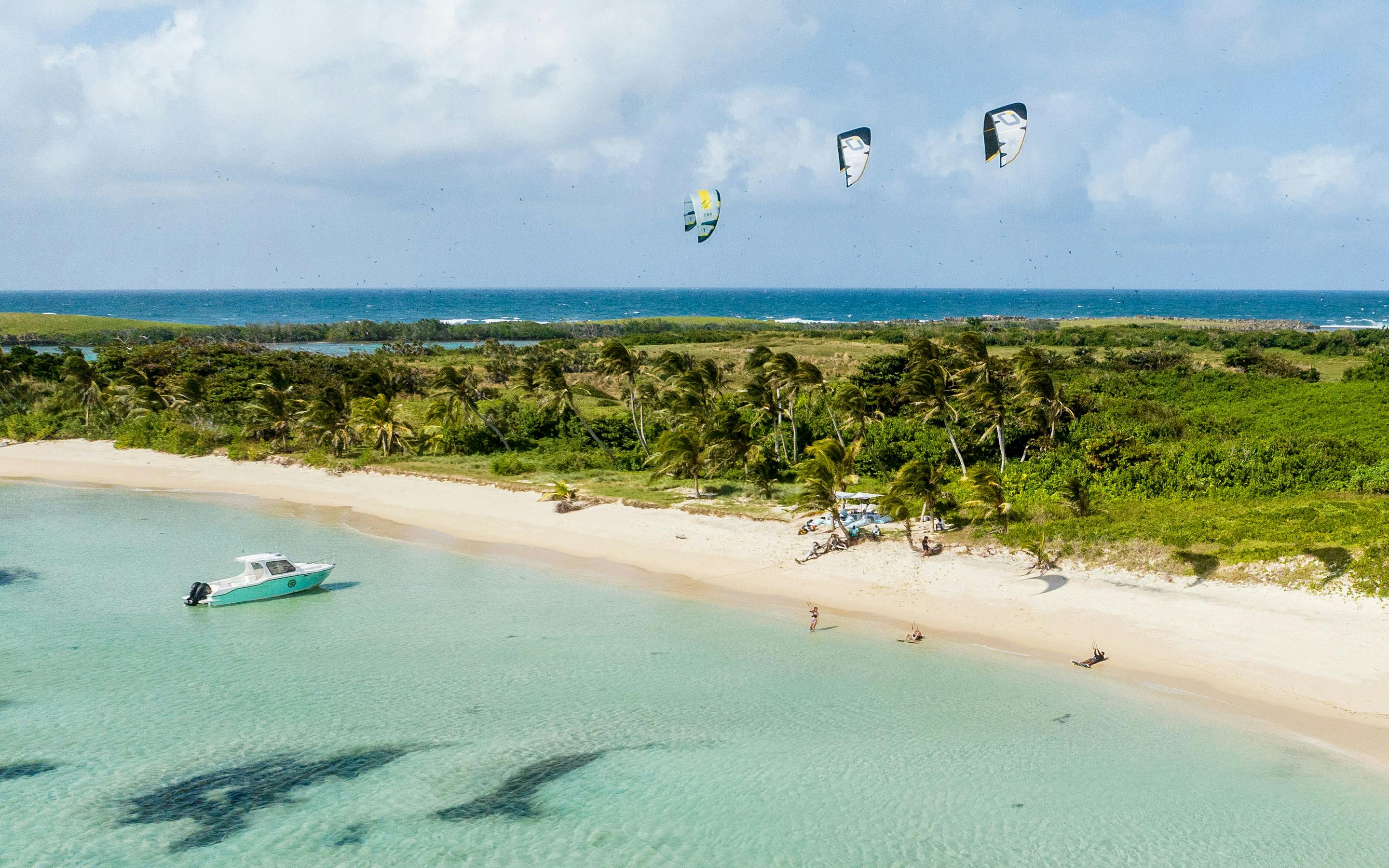 Aerial view of a white-sand beach and turquoise lagoon beside a lush island, with kitesurfers in the shallows.