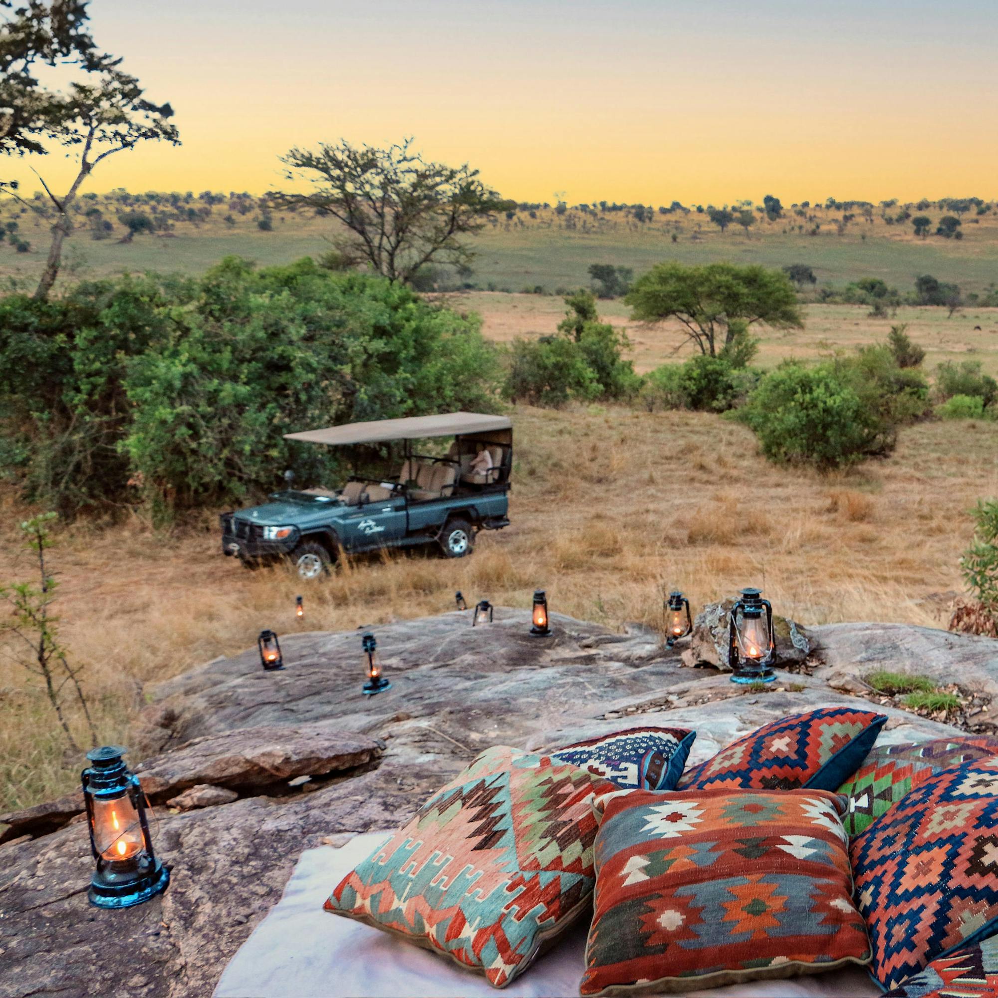 Colorful floor cushions and a low table sit on a rocky lookout at sunset, with a safari vehicle parked nearby.
