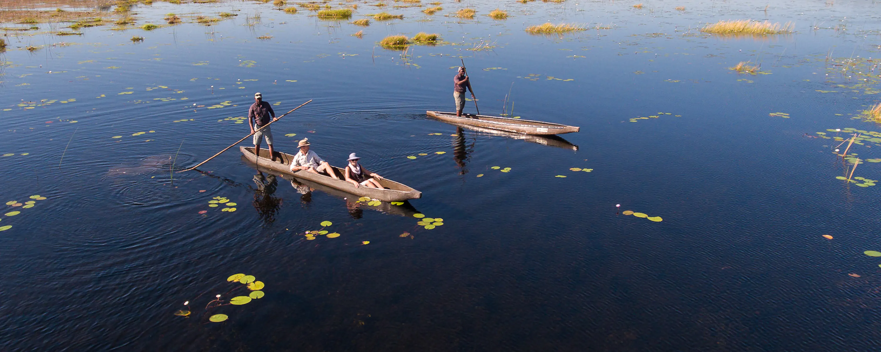 Two mokoro canoes glide over blue water dotted with lily pads, leaving soft ripples across the calm delta.