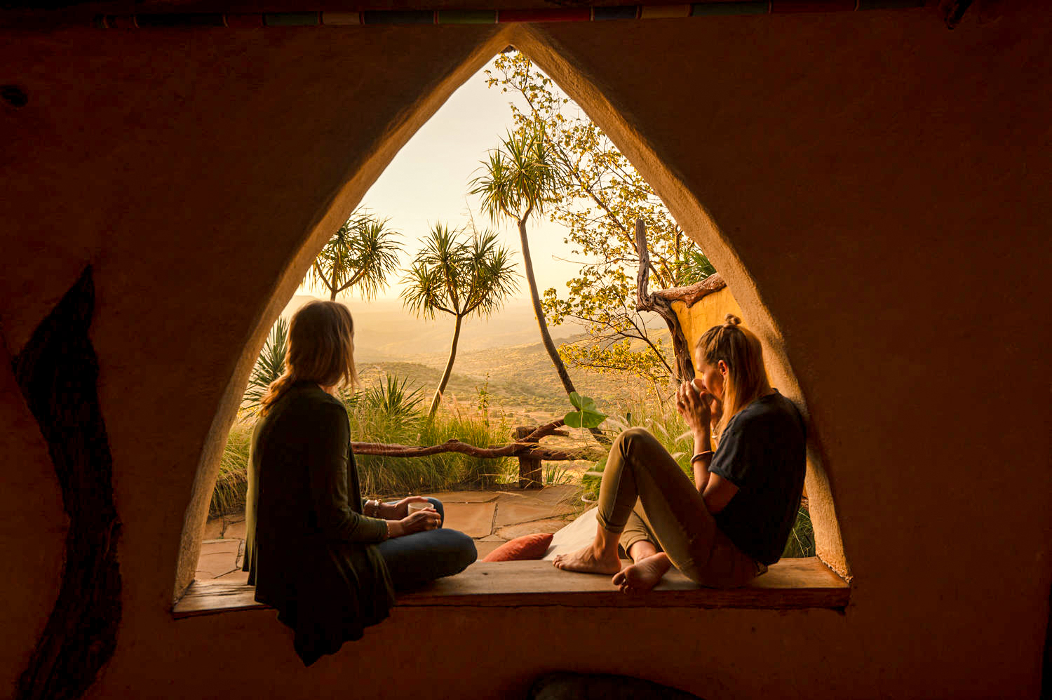Two guests relax on cushions inside a tent doorway, watching golden sunset light spill over the savannah beyond.