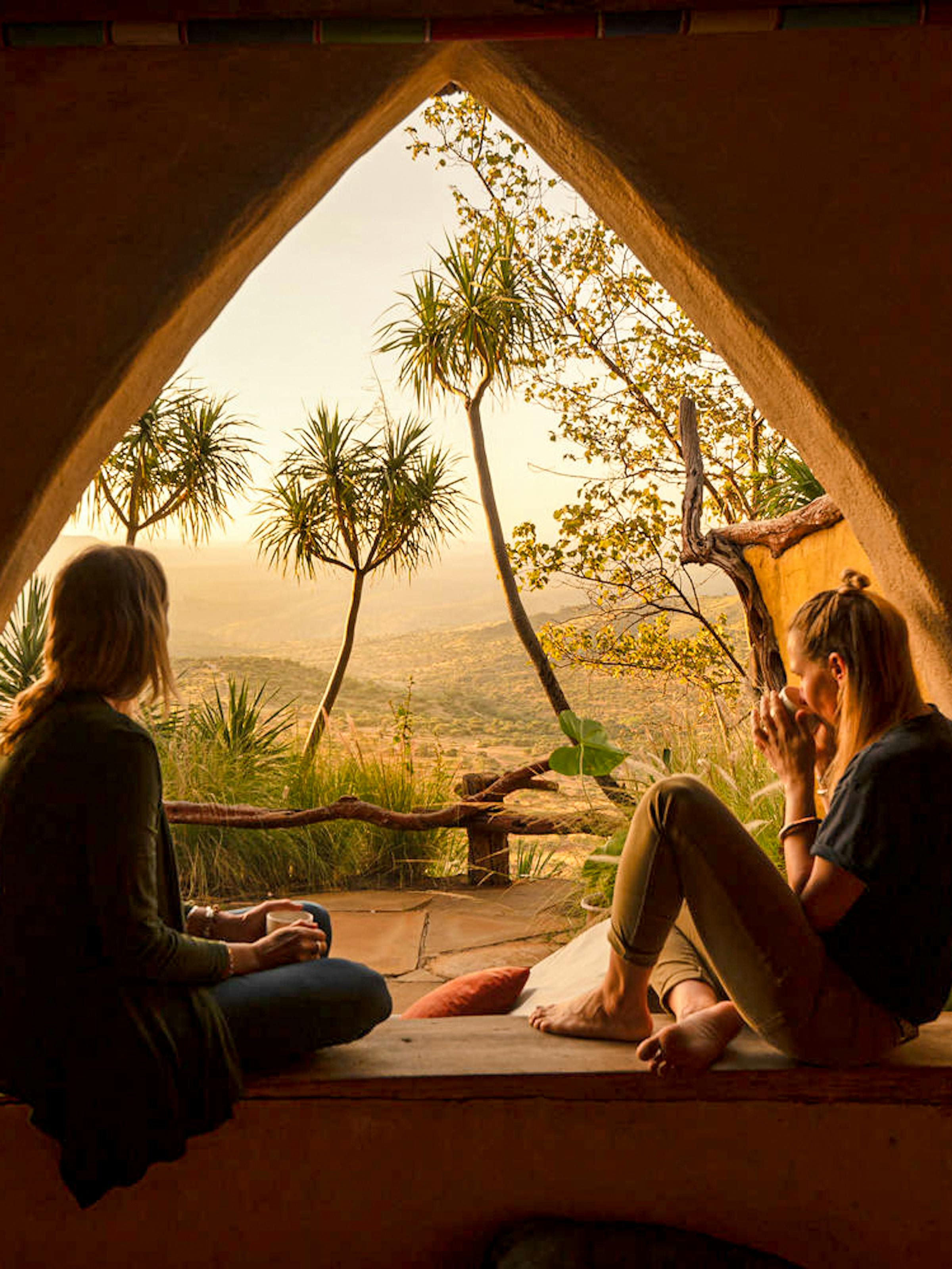 Two guests relax on cushions inside a tent doorway, watching golden sunset light spill over the savannah beyond.