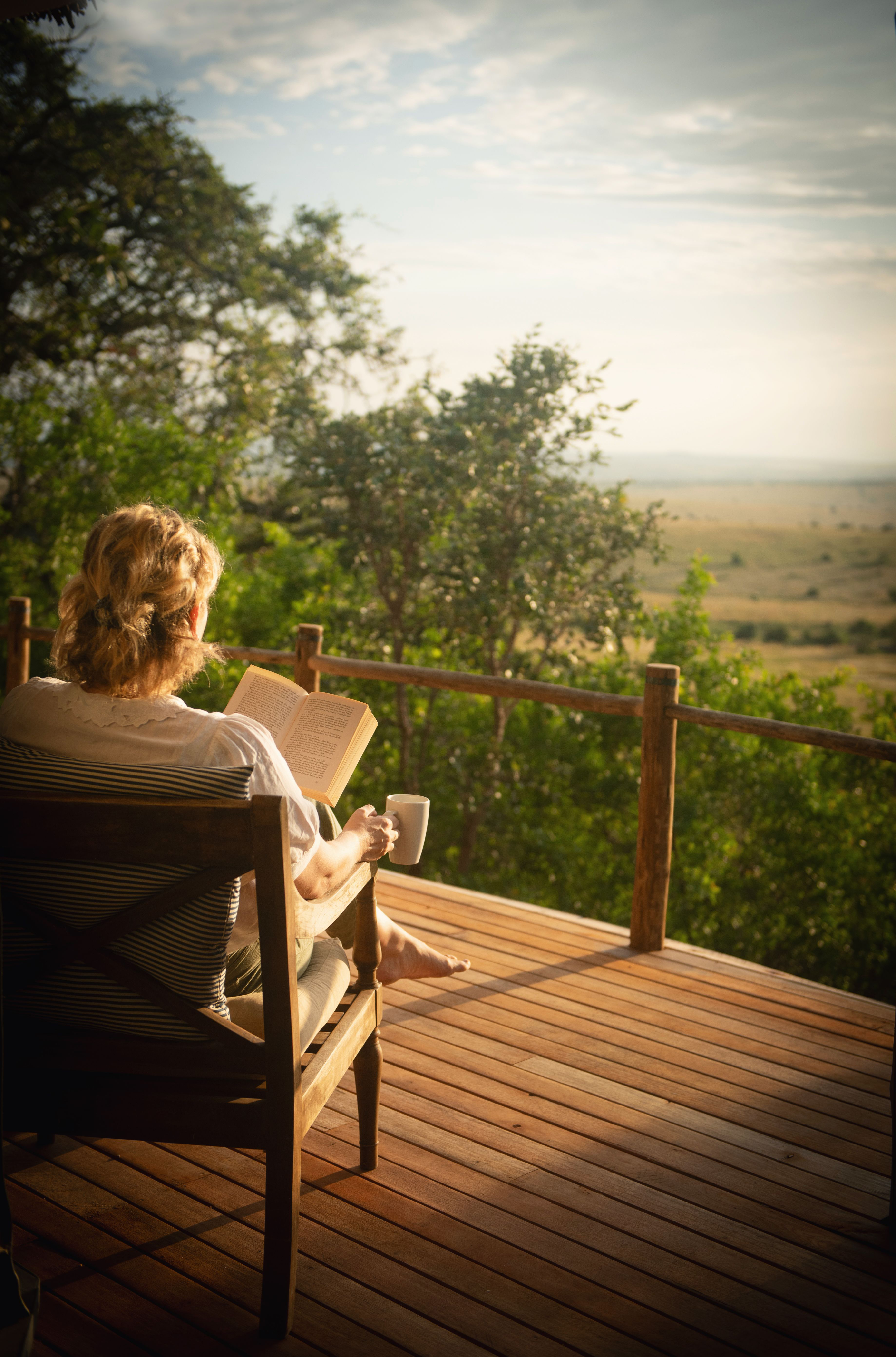 A guest sits on a lodge veranda at sunrise, looking over green plains and distant hills in soft morning light.