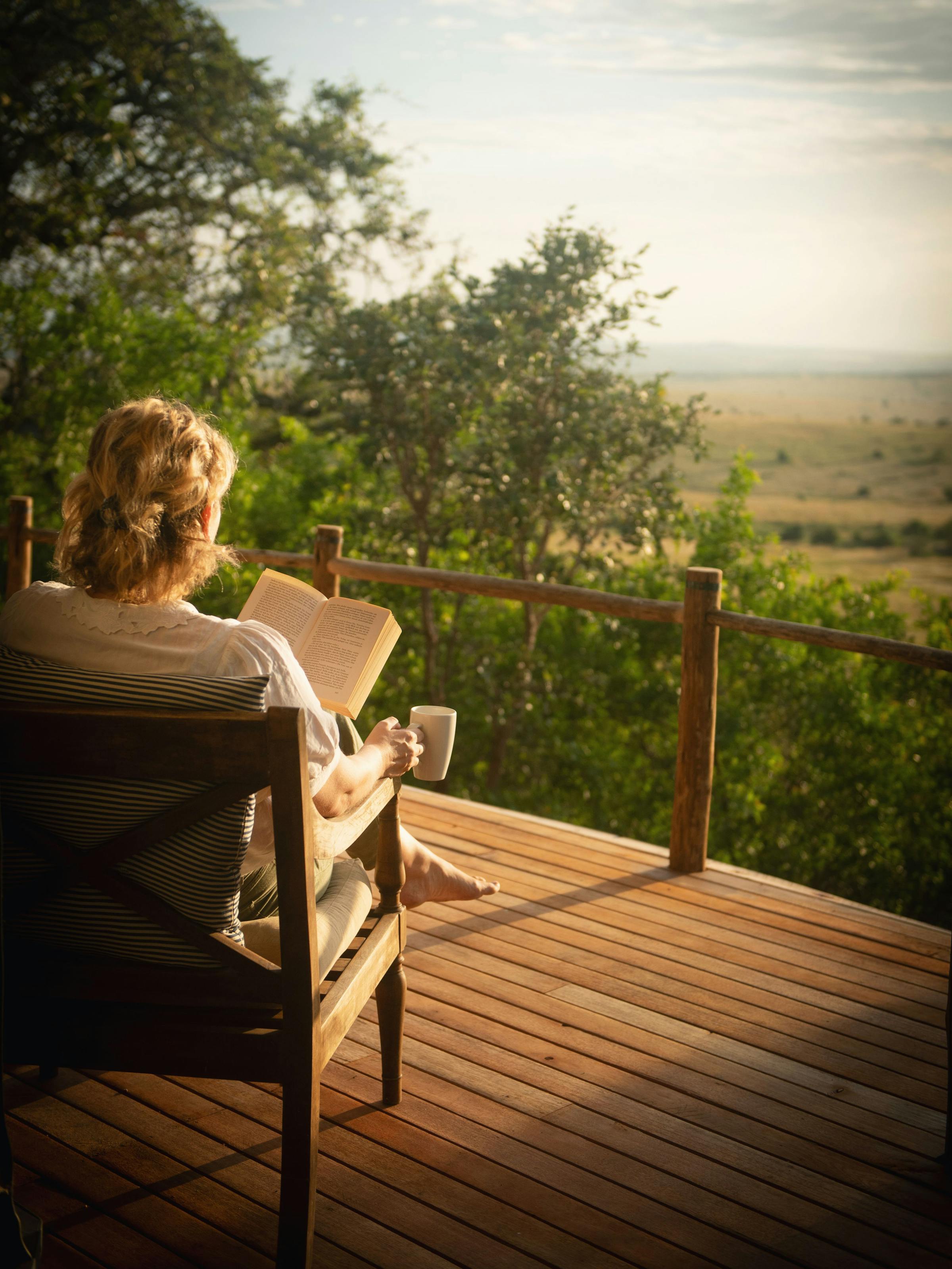 A guest sits on a lodge veranda at sunrise, looking over green plains and distant hills in soft morning light.