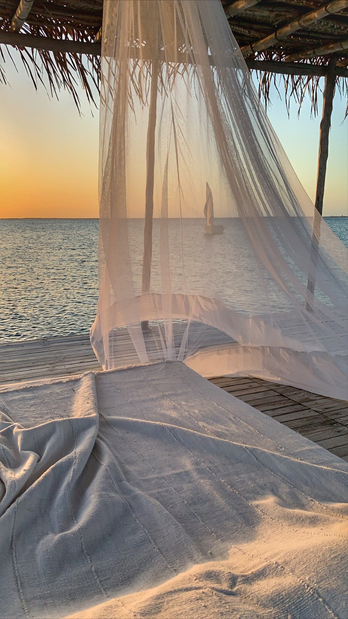 A canopy bed draped in sheer curtains faces the ocean, with warm sunset colors glowing above a calm horizon.