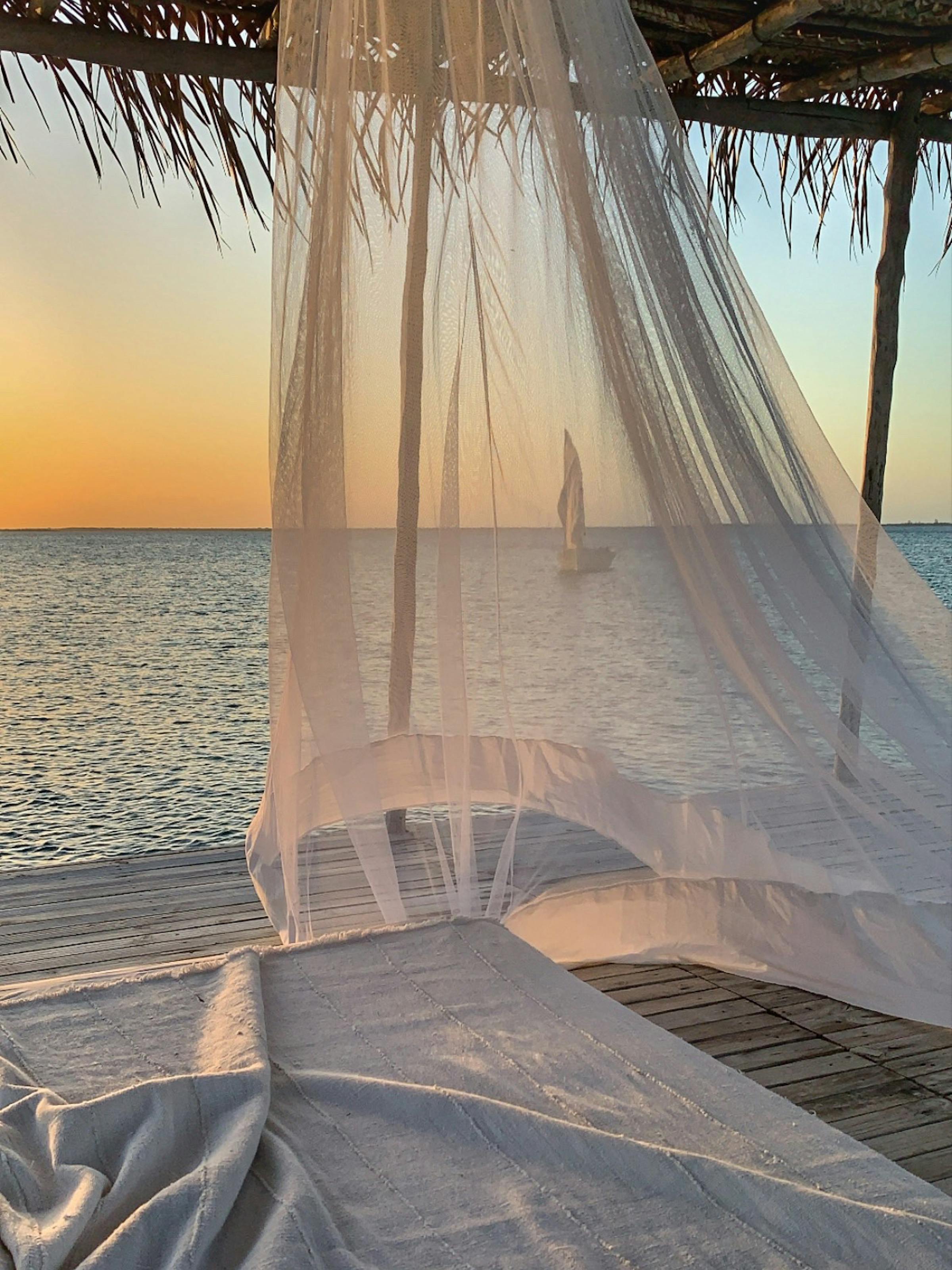 A canopy bed draped in sheer curtains faces the ocean, with warm sunset colors glowing above a calm horizon.
