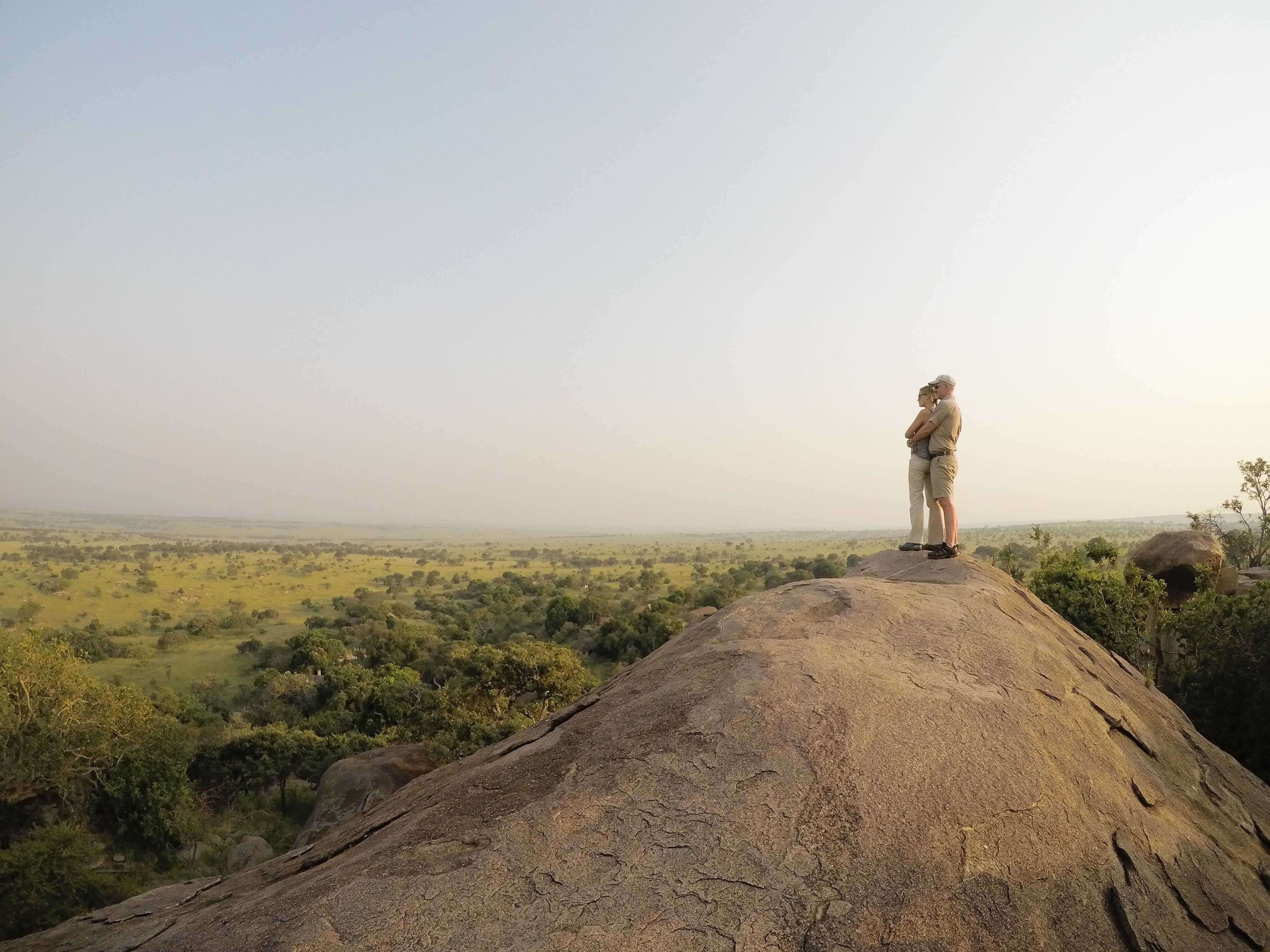 A lone figure stands on a rocky outcrop, looking across hazy plains stretching into the quiet distance below.
