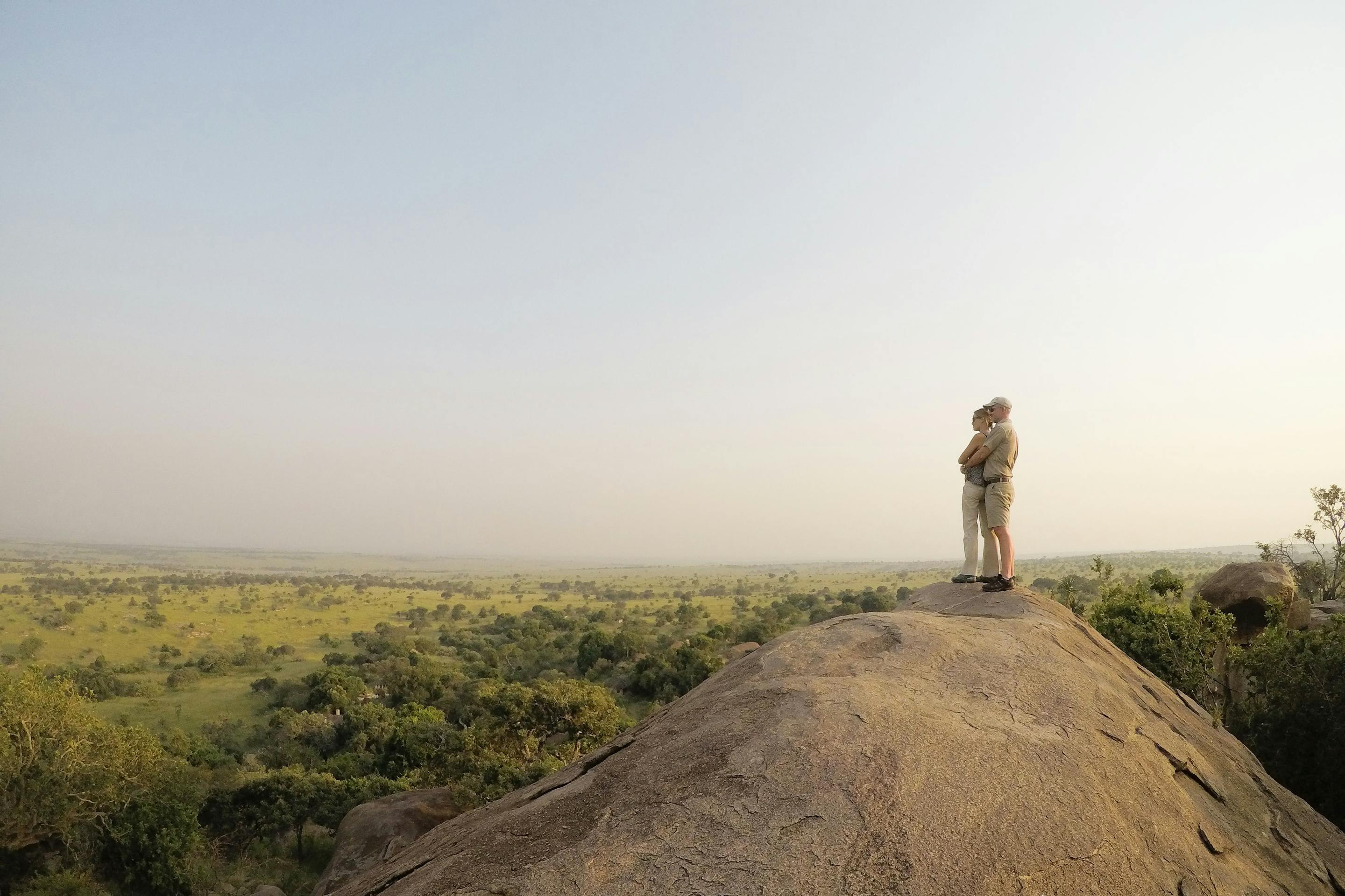 A lone figure stands on a rocky outcrop, looking across hazy plains stretching into the quiet distance below.