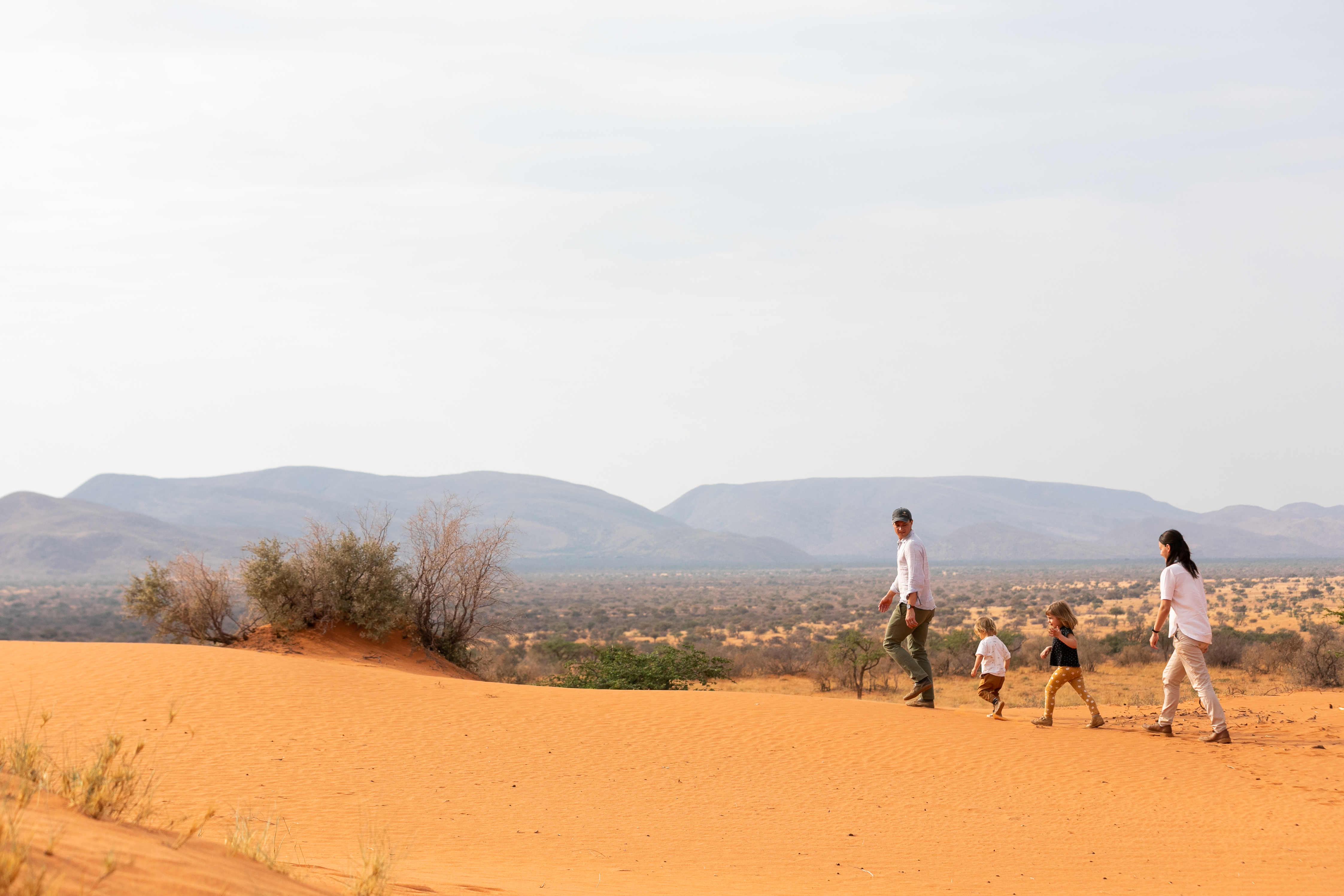 A small group walks across red sand dunes toward distant mountains, leaving a trail of footprints behind.