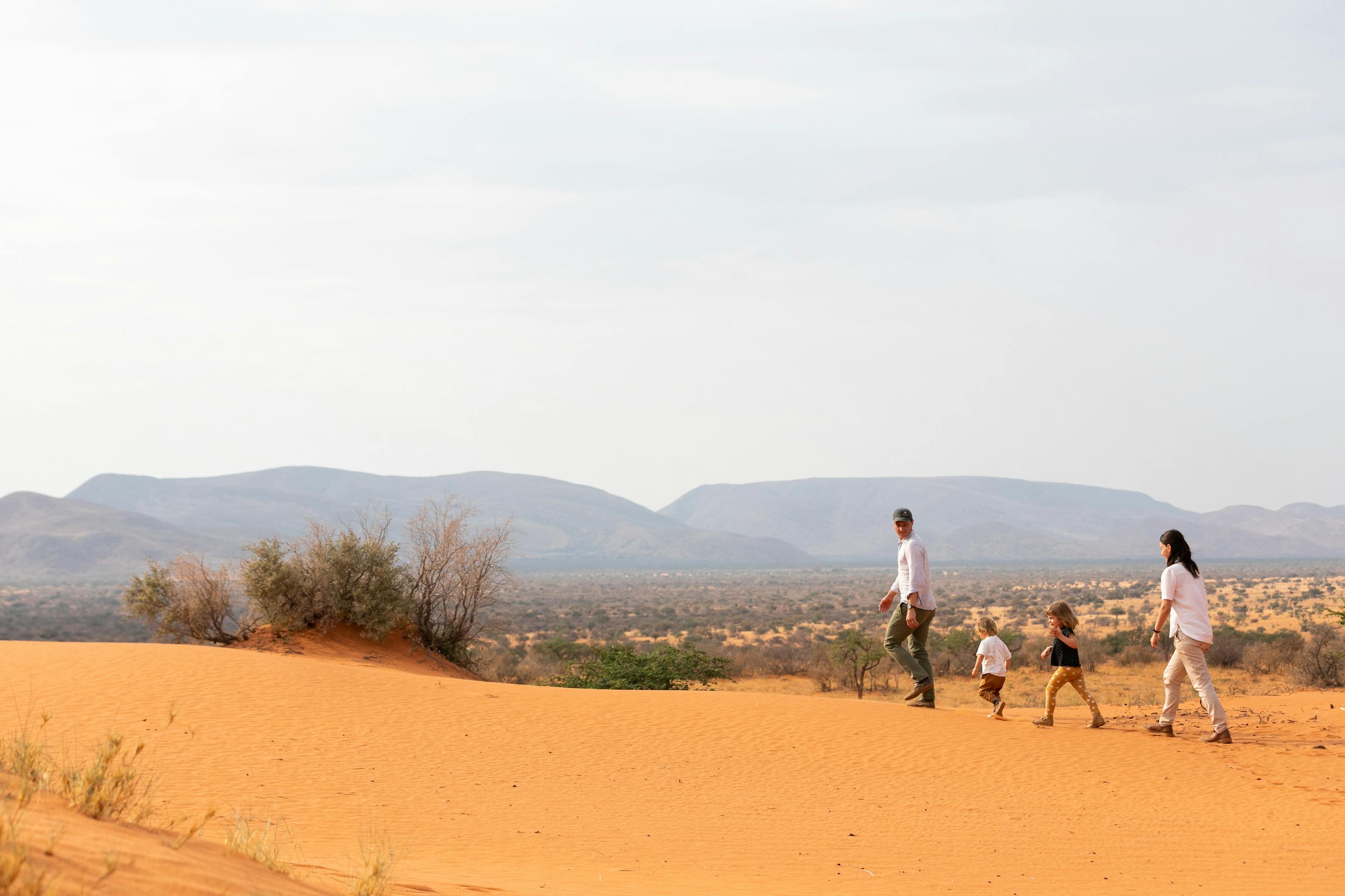 A small group walks across red sand dunes toward distant mountains, leaving a trail of footprints behind.