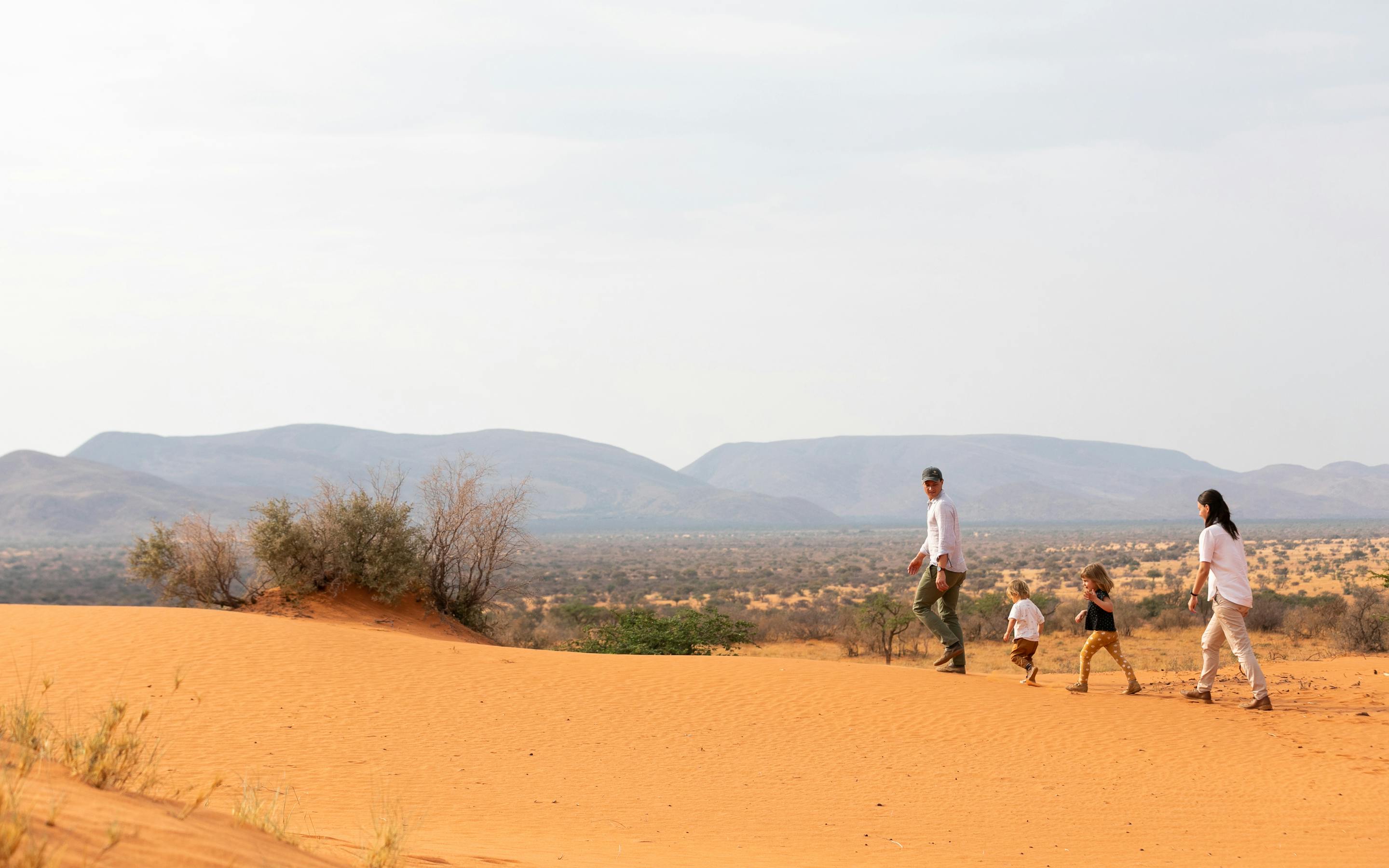 A small group walks across red sand dunes toward distant mountains, leaving a trail of footprints behind.