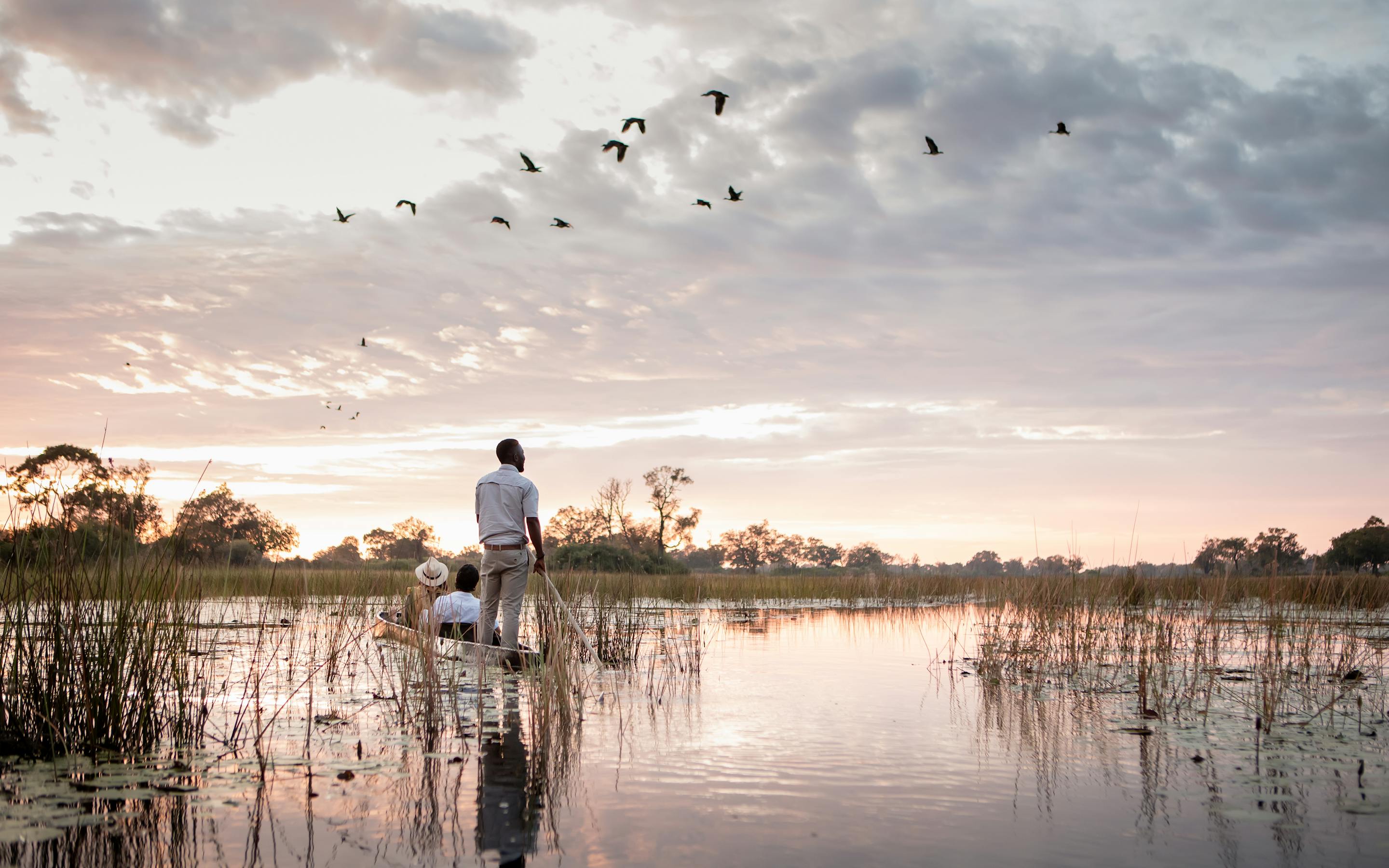 A person poles a canoe through shallow reflective water at dusk, as birds circle above and reeds frame the scene.
