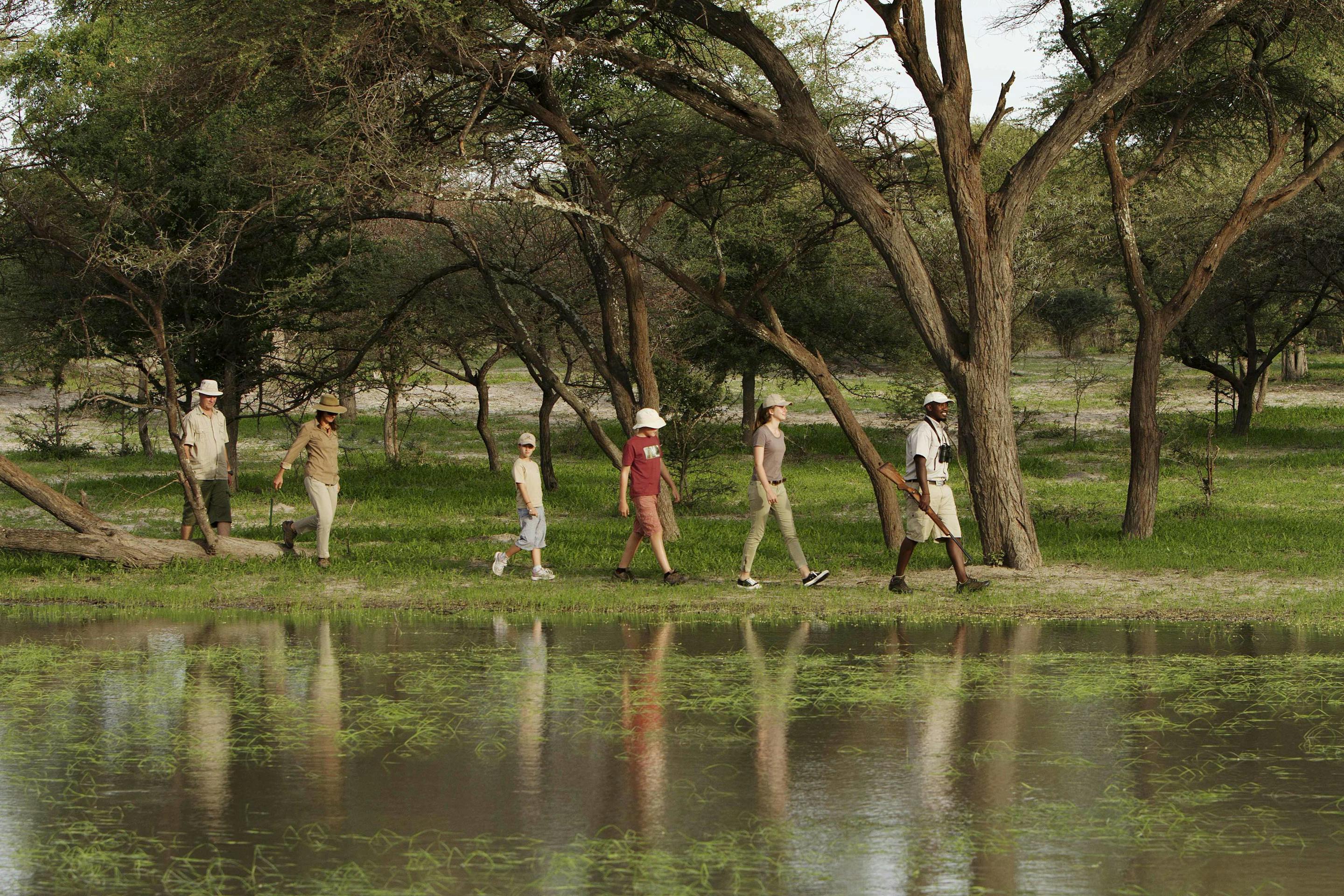 A small group walks single file through shallow floodwater beneath trees, with reflections shimmering around them.