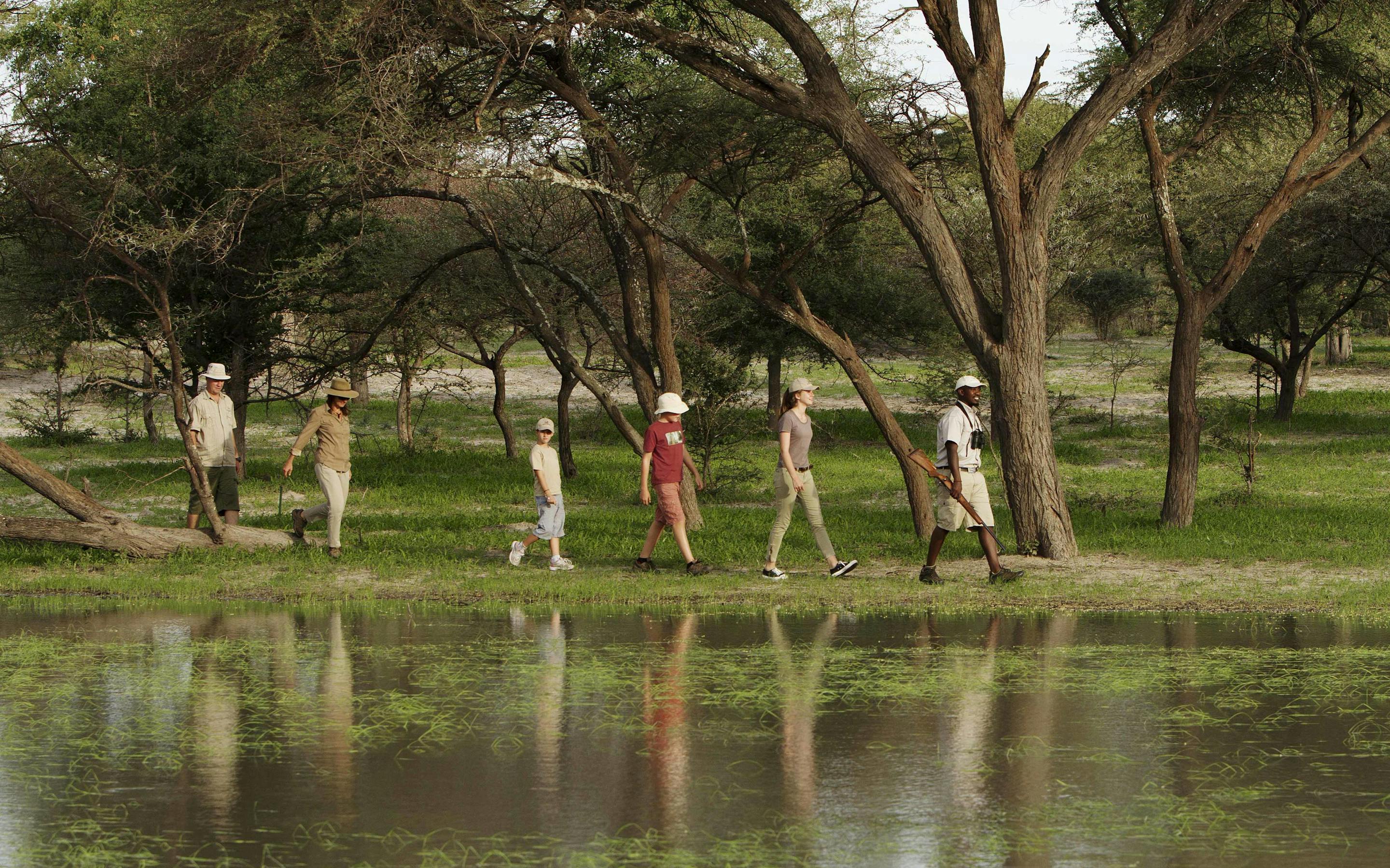 A small group walks single file through shallow floodwater beneath trees, with reflections shimmering around them.
