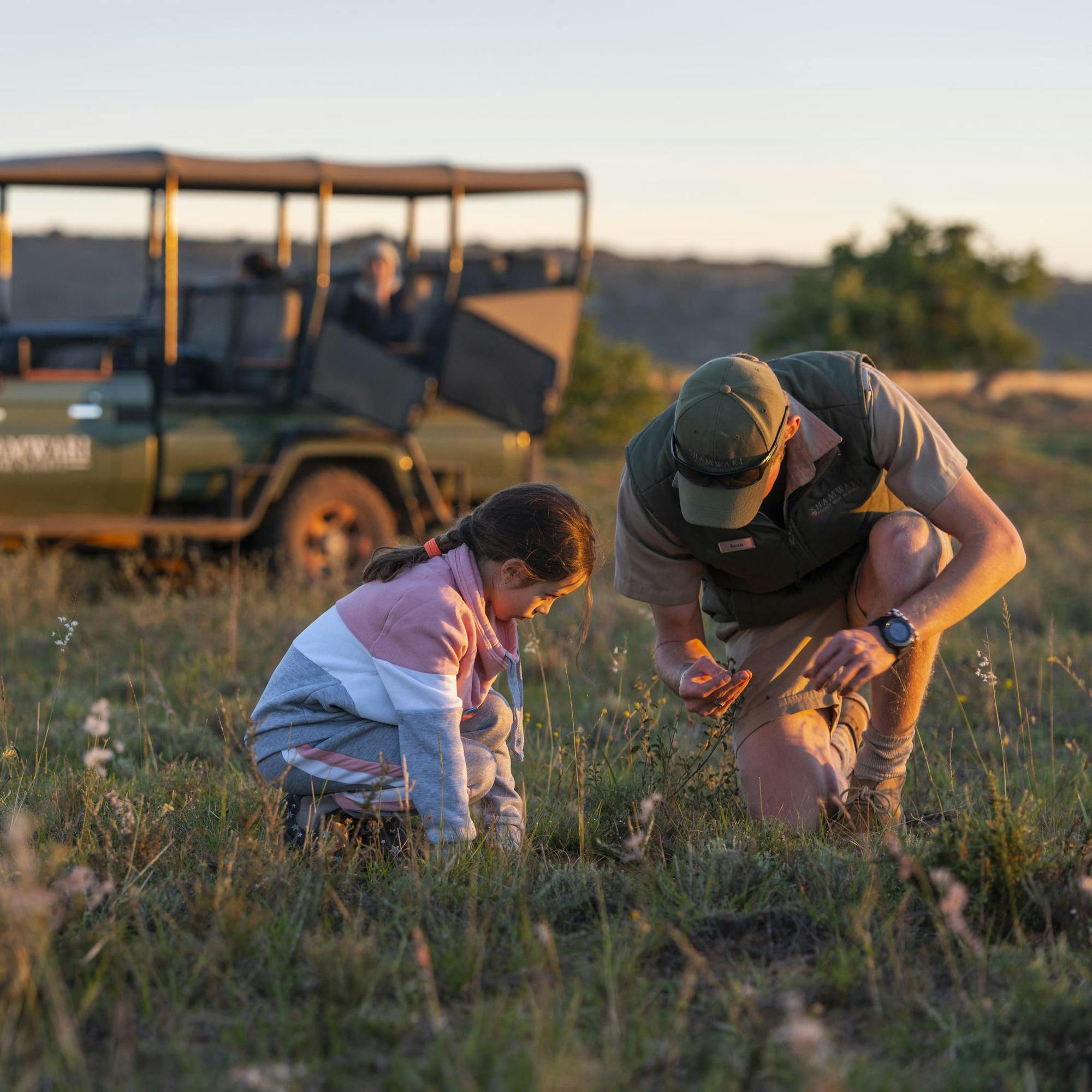 Two children sit in an open safari vehicle, looking out over grassland as wildlife grazes in the distance.