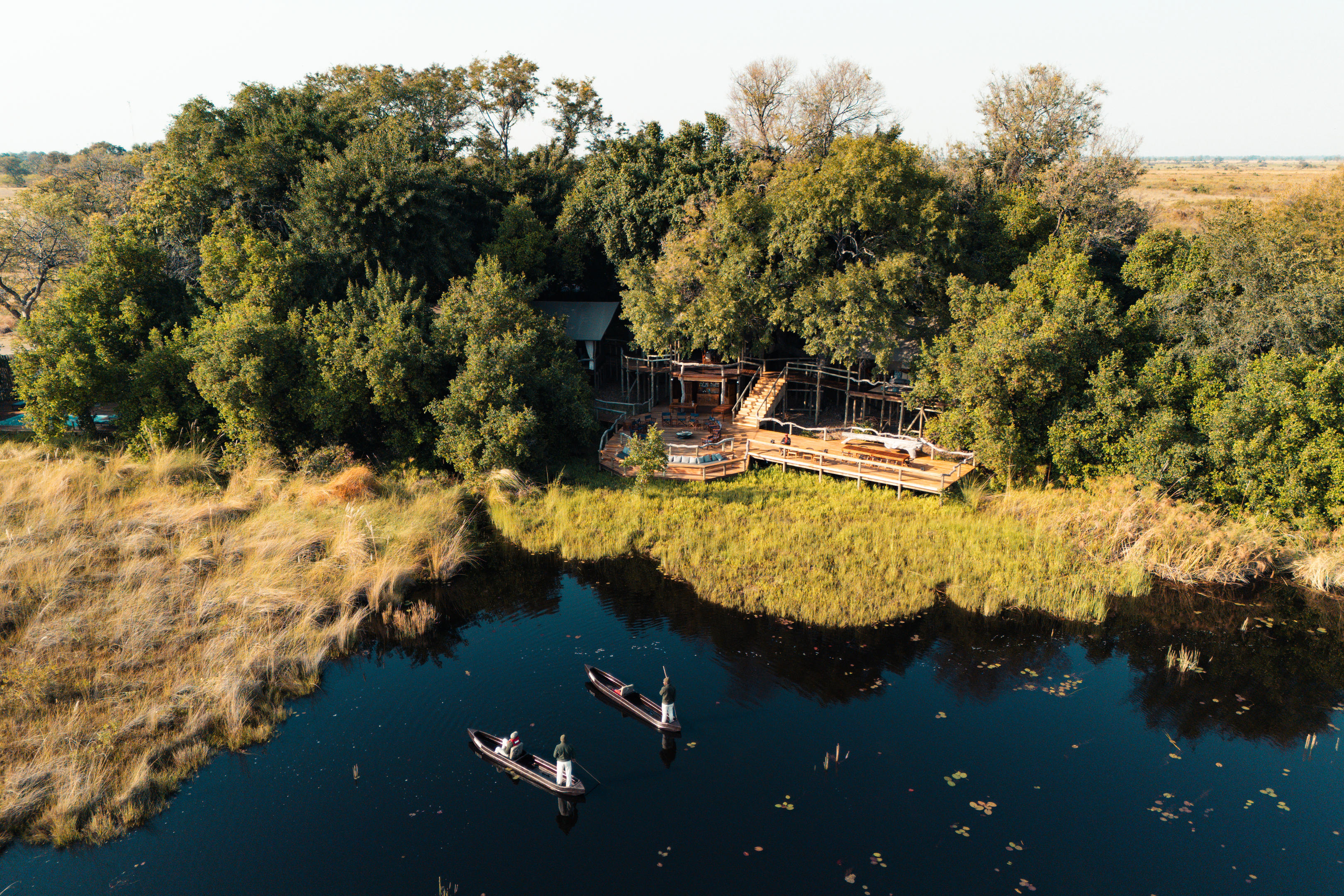 Aerial view of a riverside lodge in dense trees, with a wooden deck and two canoes on calm dark water.