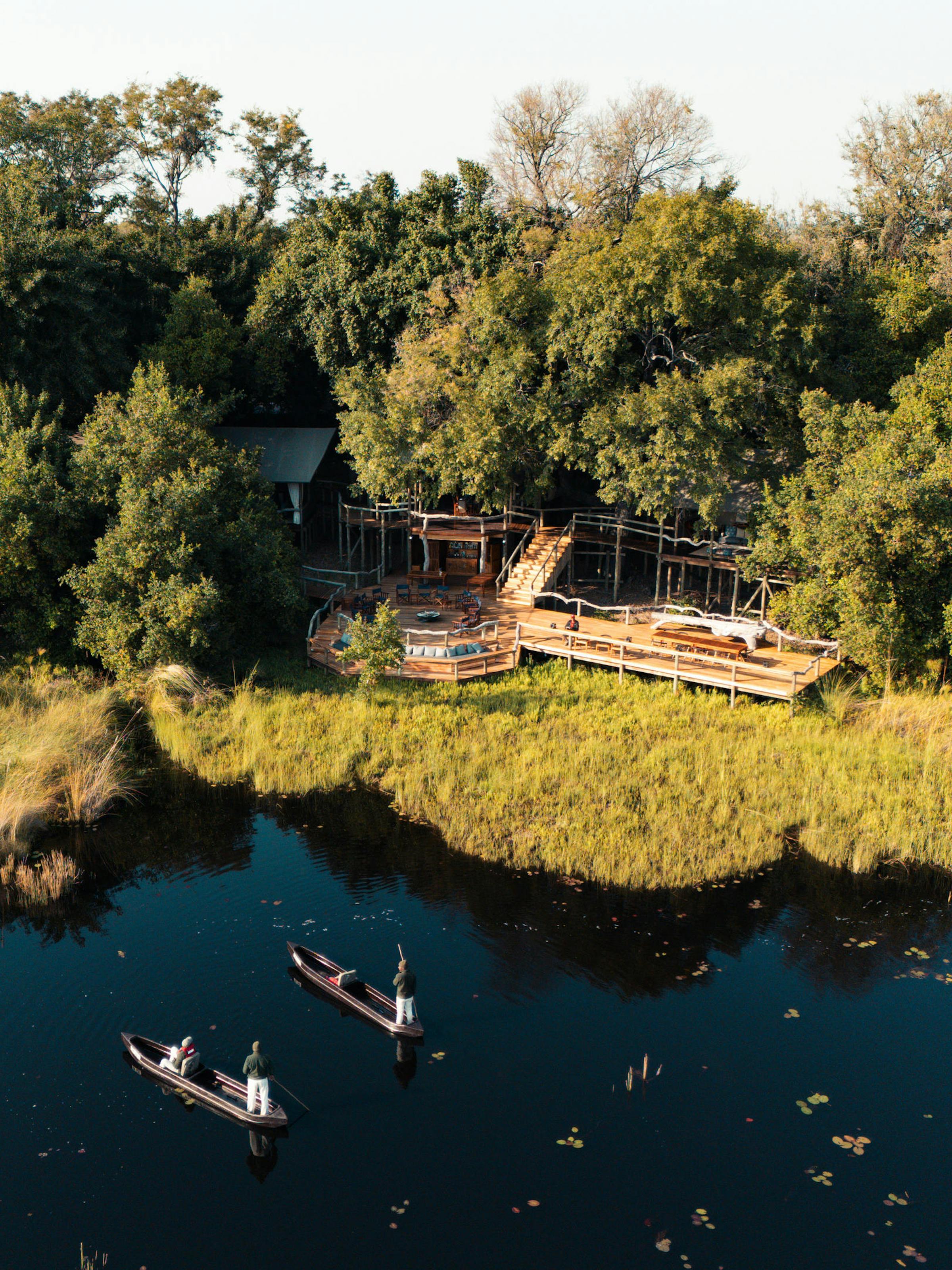 Aerial view of a riverside lodge in dense trees, with a wooden deck and two canoes on calm dark water.