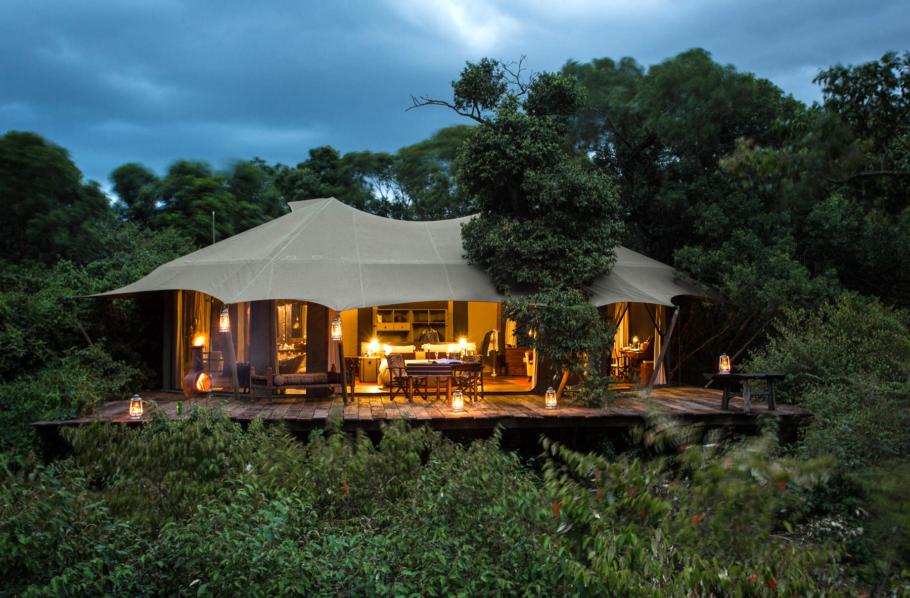 Tented safari suite glows at night with lantern light, set on a wooden deck with chairs and a covered roof.