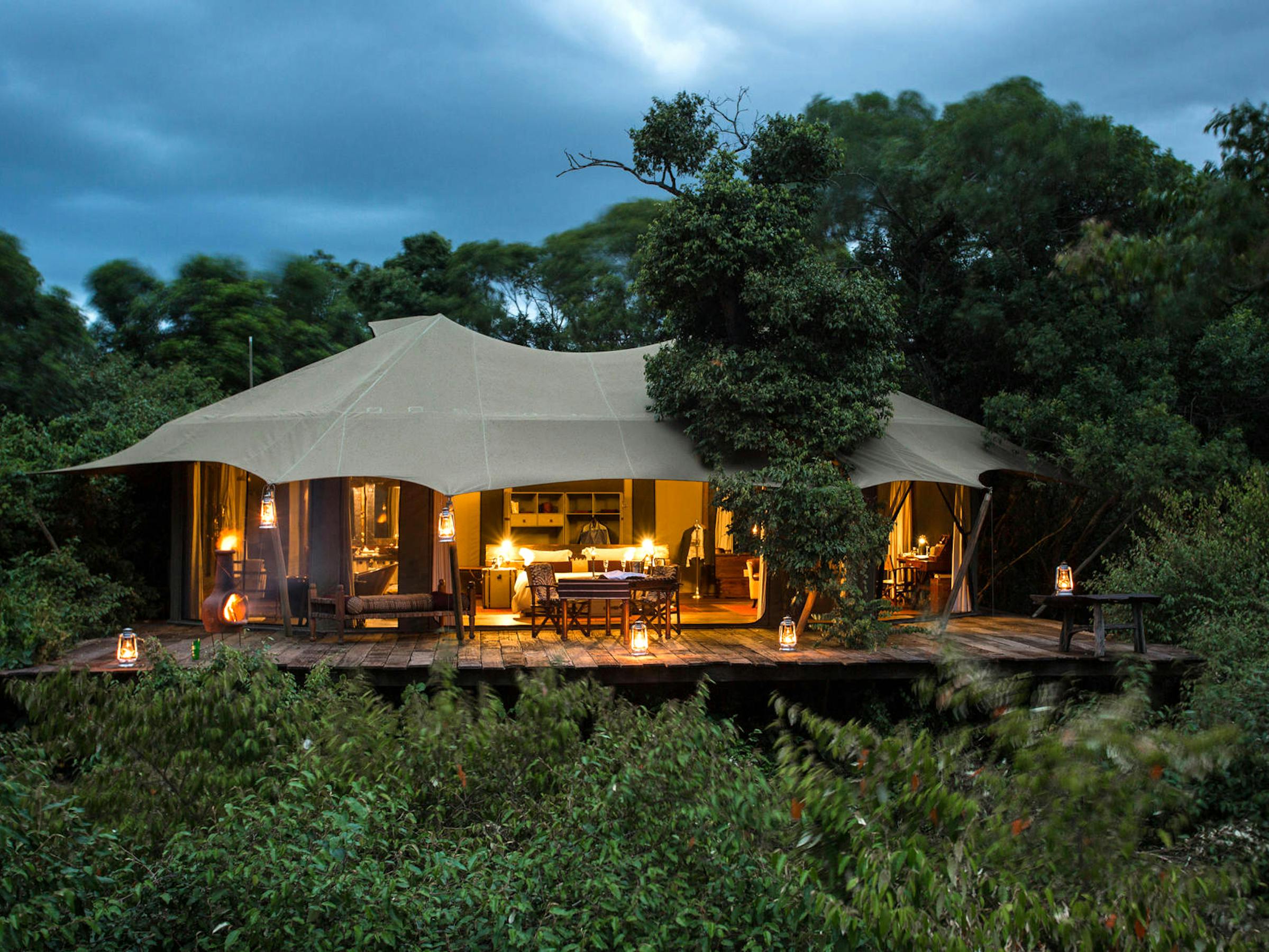 Tented safari suite glows at night with lantern light, set on a wooden deck with chairs and a covered roof.