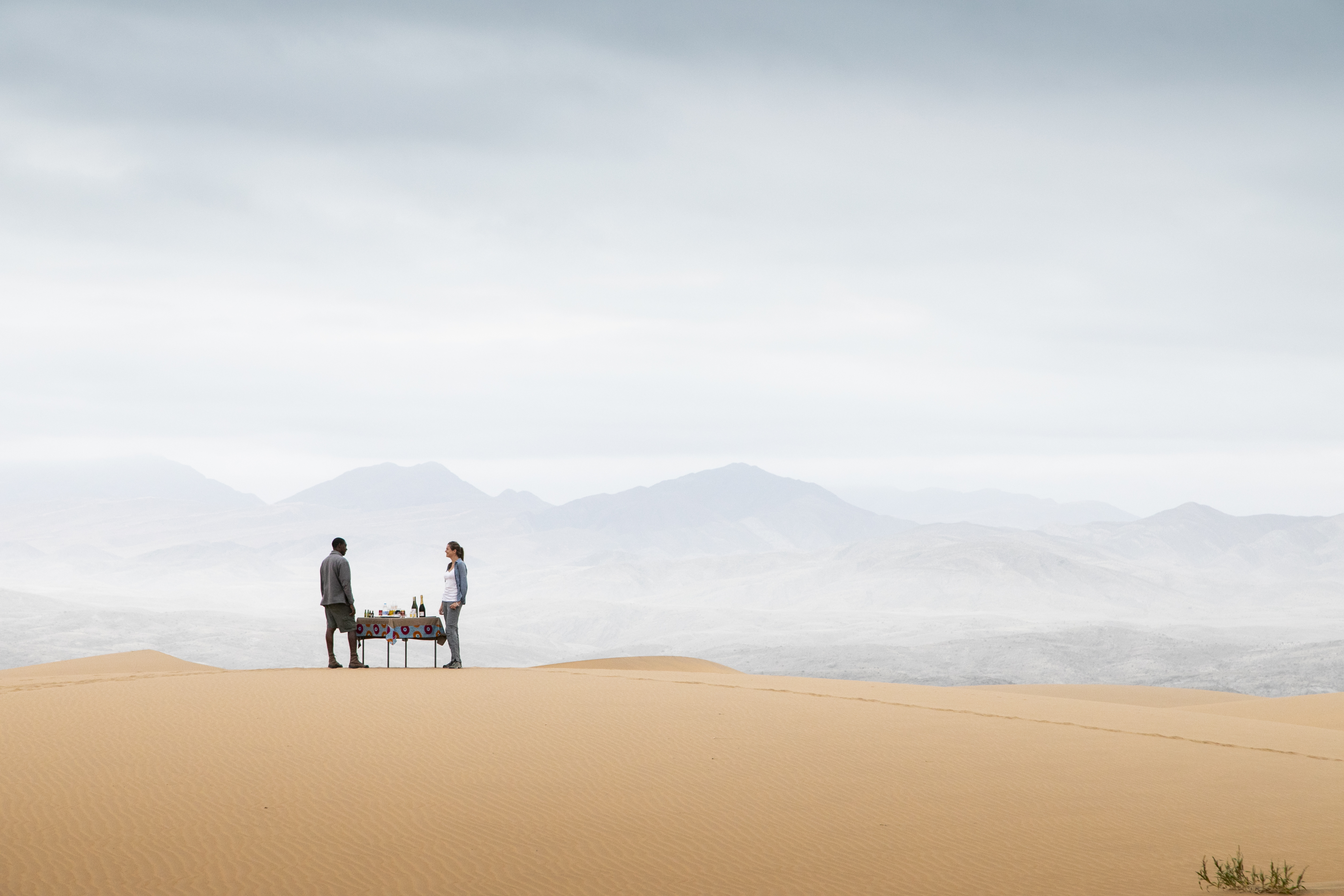 Two people stand on a high sand dune overlooking rippled desert slopes, with long shadows and clear sky.