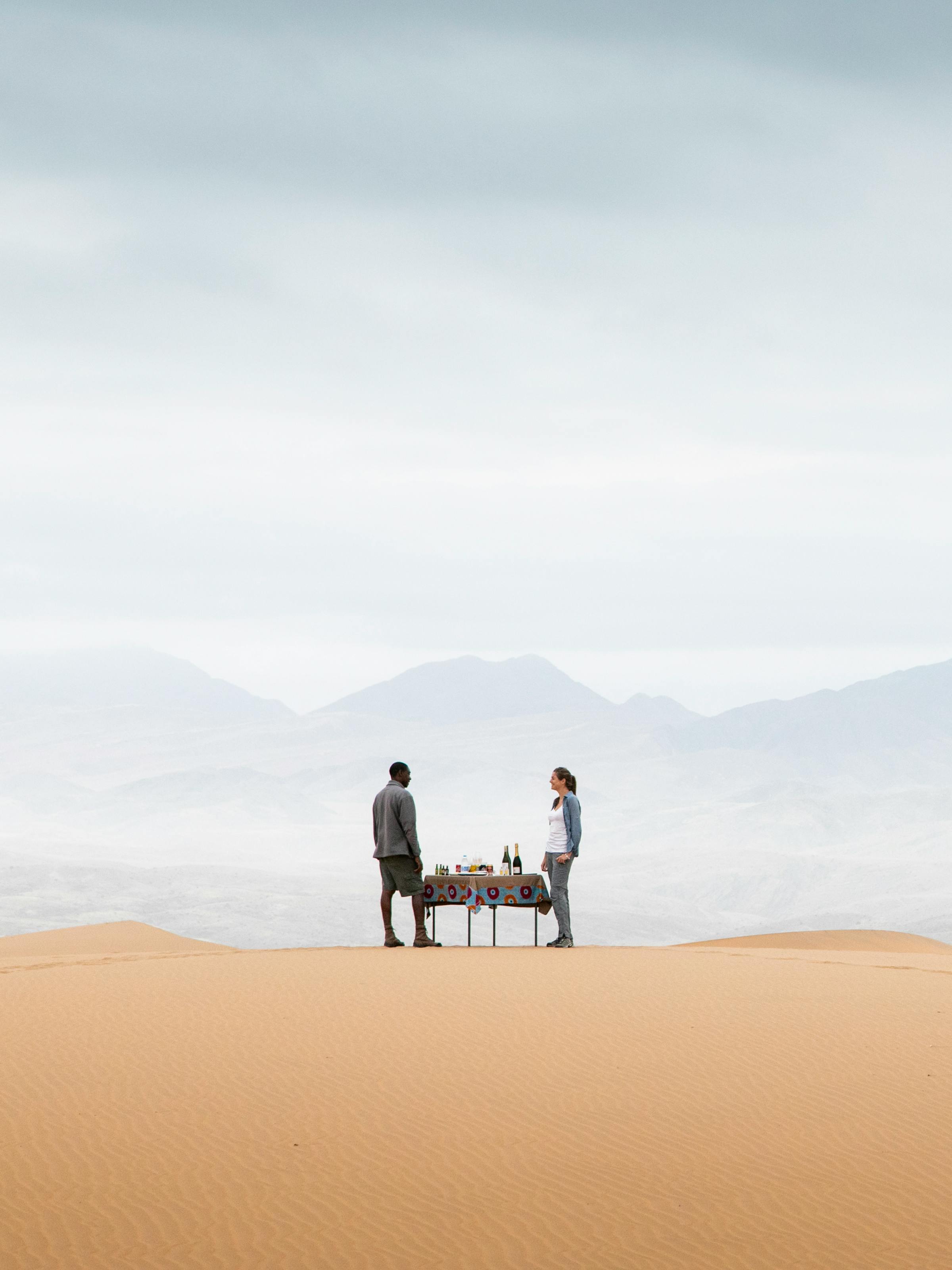 Two people stand on a high sand dune overlooking rippled desert slopes, with long shadows and clear sky.