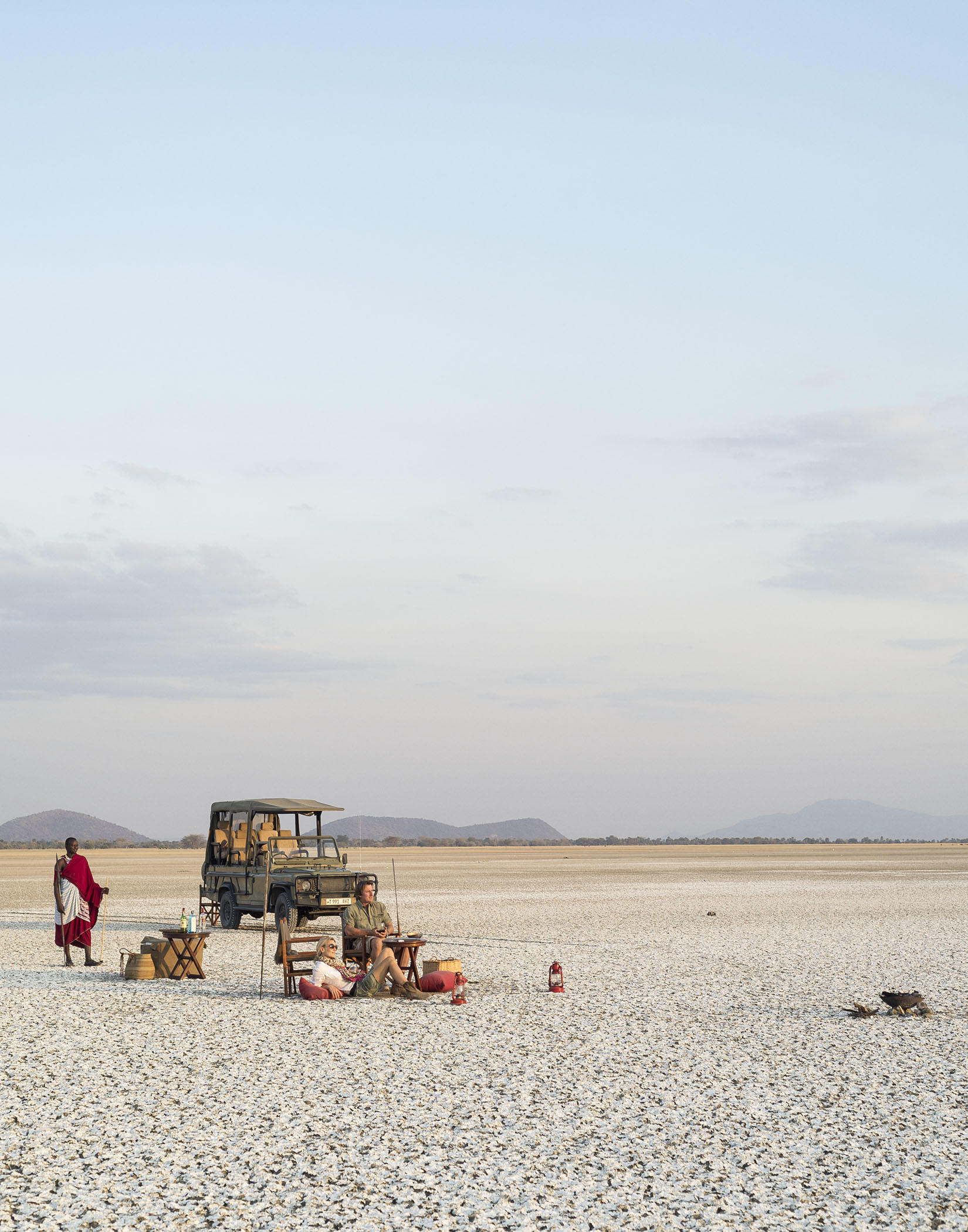 Safari vehicle and picnic chairs sit on a vast white salt pan beneath a pale sunset sky and distant hills.
