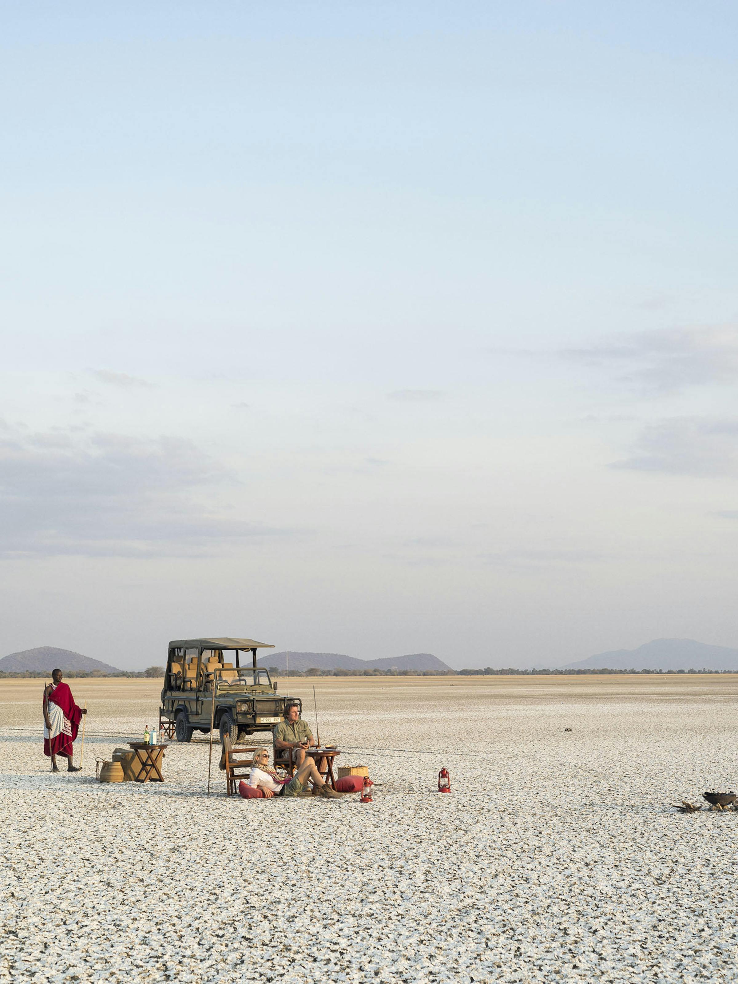 Safari vehicle and picnic chairs sit on a vast white salt pan beneath a pale sunset sky and distant hills.