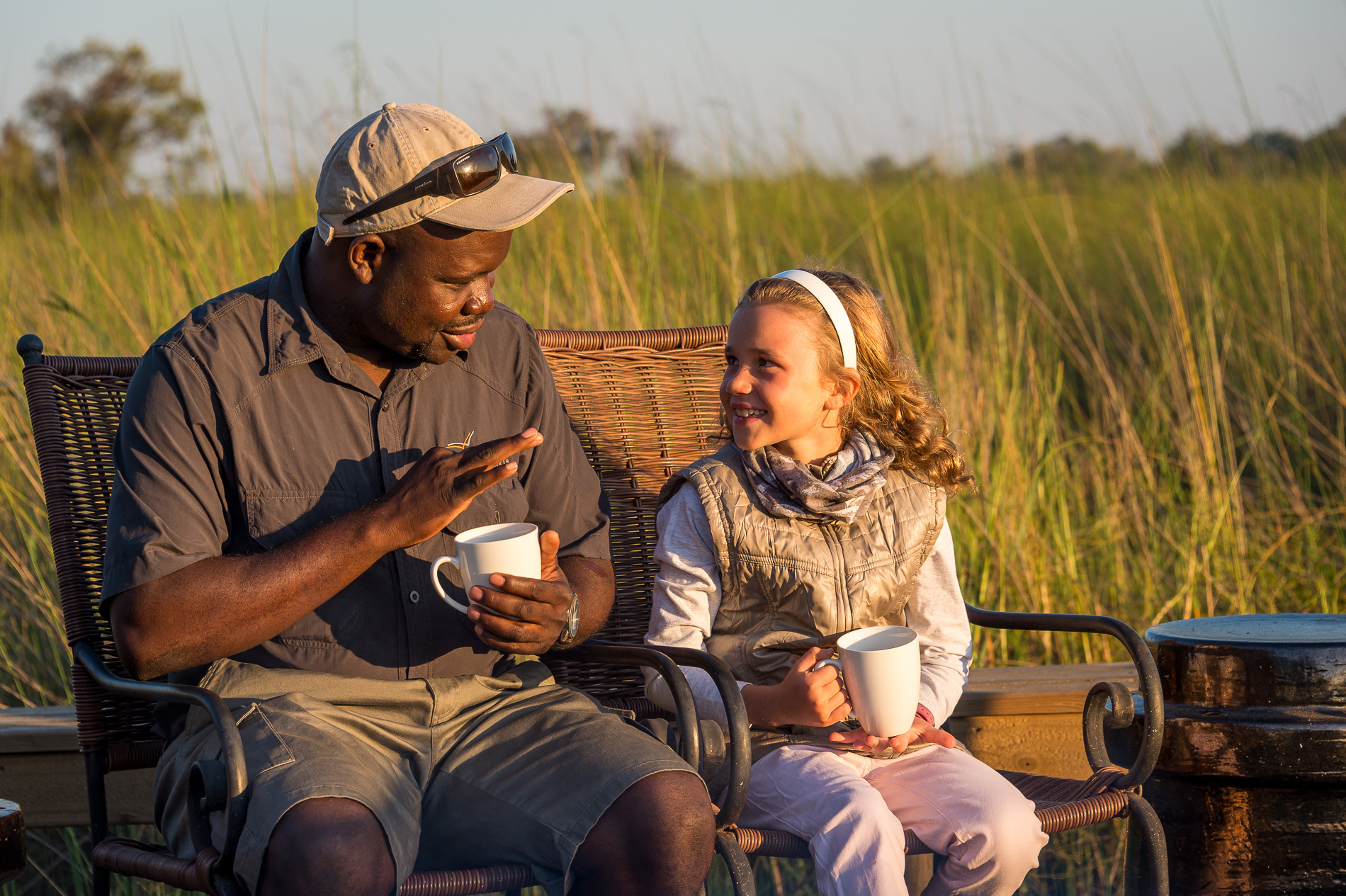 Two people sit in a safari boat in tall reeds, sharing warm drinks as golden light settles on still water.