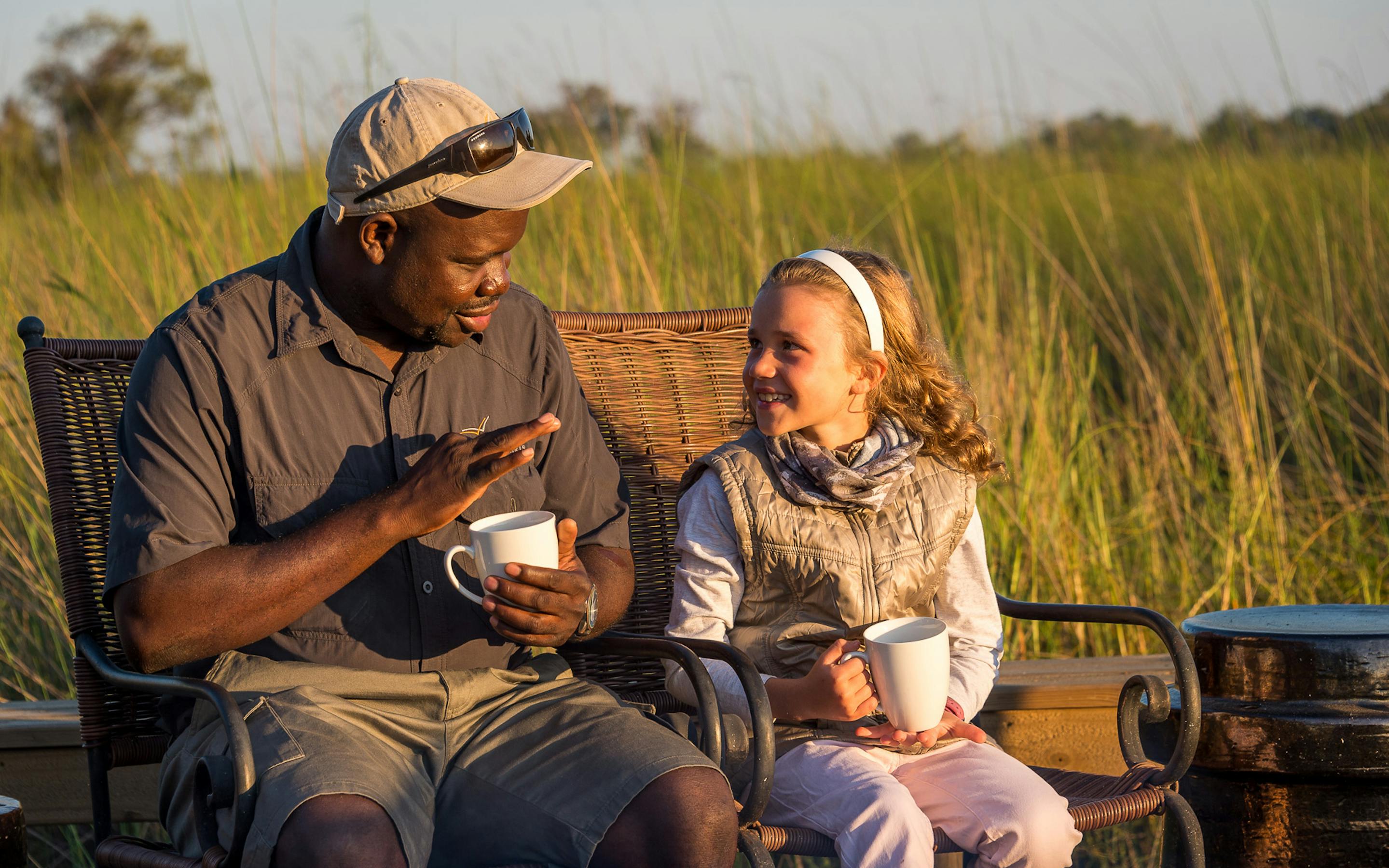 Two people sit in a safari boat in tall reeds, sharing warm drinks as golden light settles on still water.
