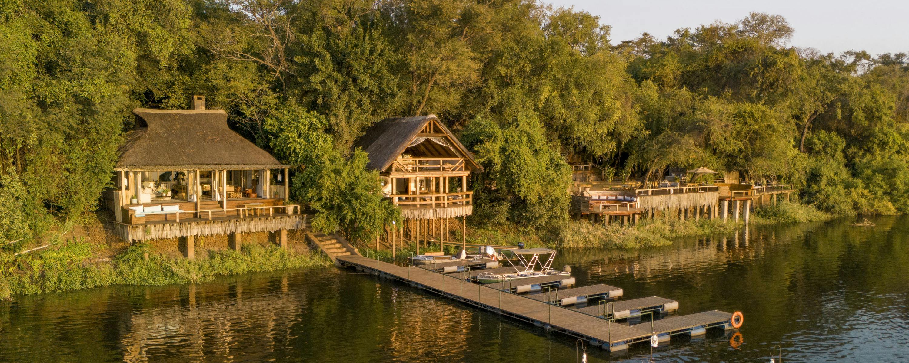 Riverside lodge suites overlook calm water, with a wooden deck and a small boat moored along the green riverbank.