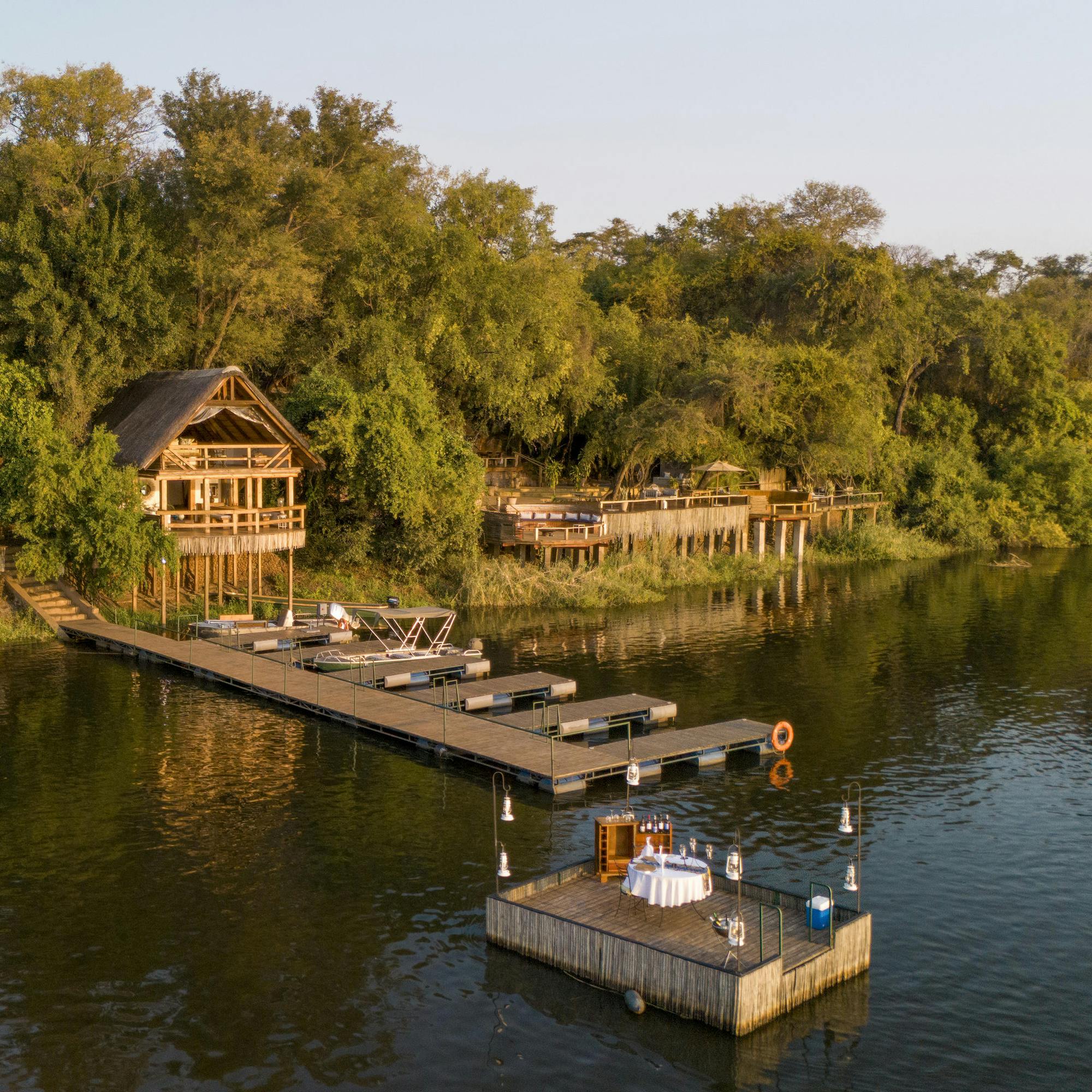 Riverside lodge suites overlook calm water, with a wooden deck and a small boat moored along the green riverbank.