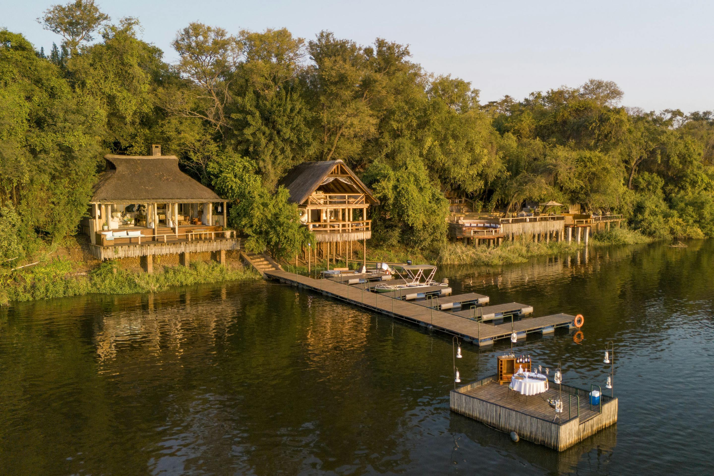 Riverside lodge suites overlook calm water, with a wooden deck and a small boat moored along the green riverbank.