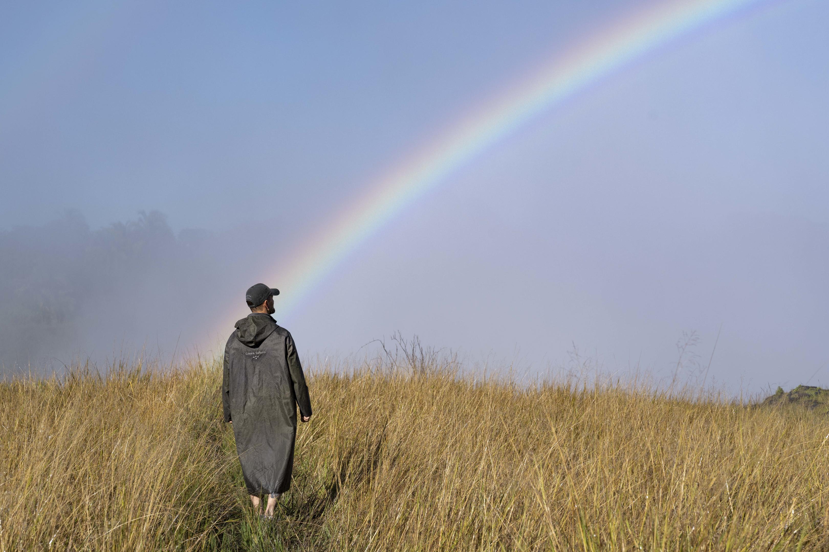 A traveler stands in tall golden grass as a bright rainbow arcs across a misty blue sky after the rain nearby.